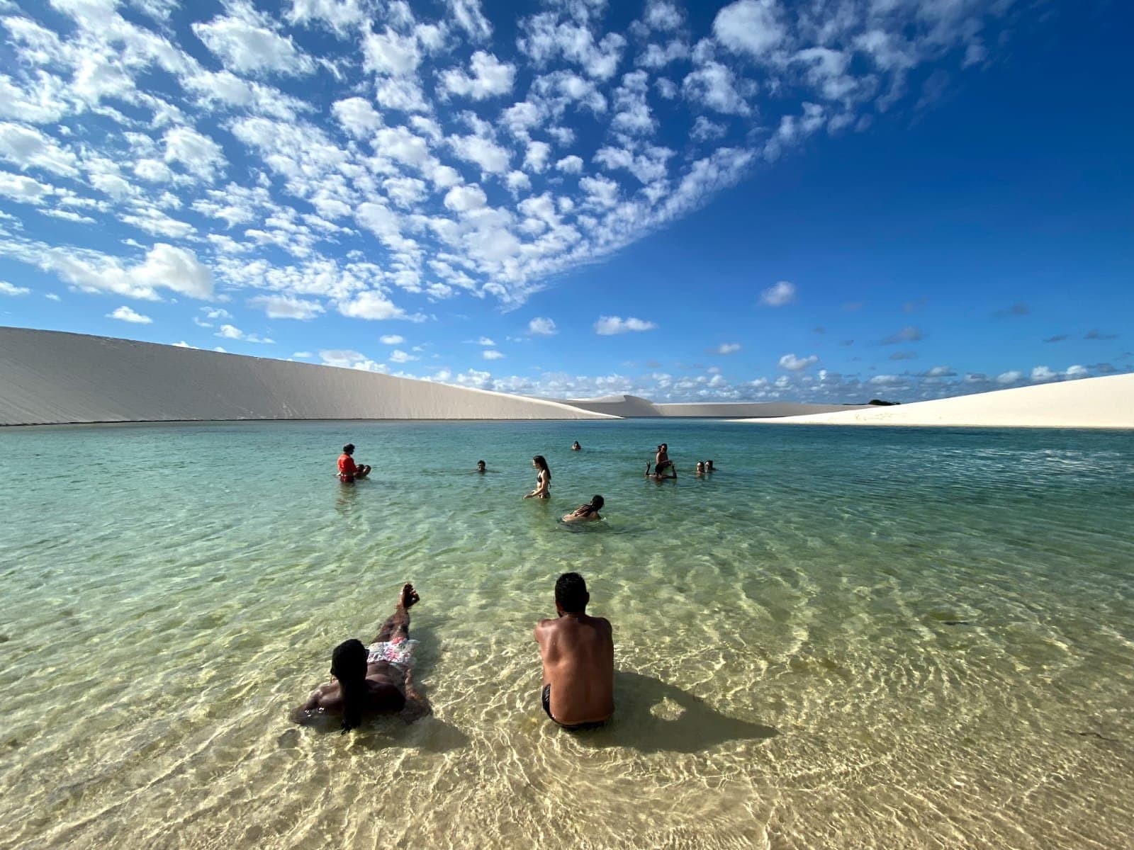 Fraîcheur de l'eau, chaleur du sable — la pause parfaite au cœur des dunes. Un groupe de voyageurs se baigne et se détend dans une lagune d'eau douce cristalline lors d'une randonnée dans les Lençóis Maranhenses, encadrée de dunes de sable blanc sous un ciel bleu vif