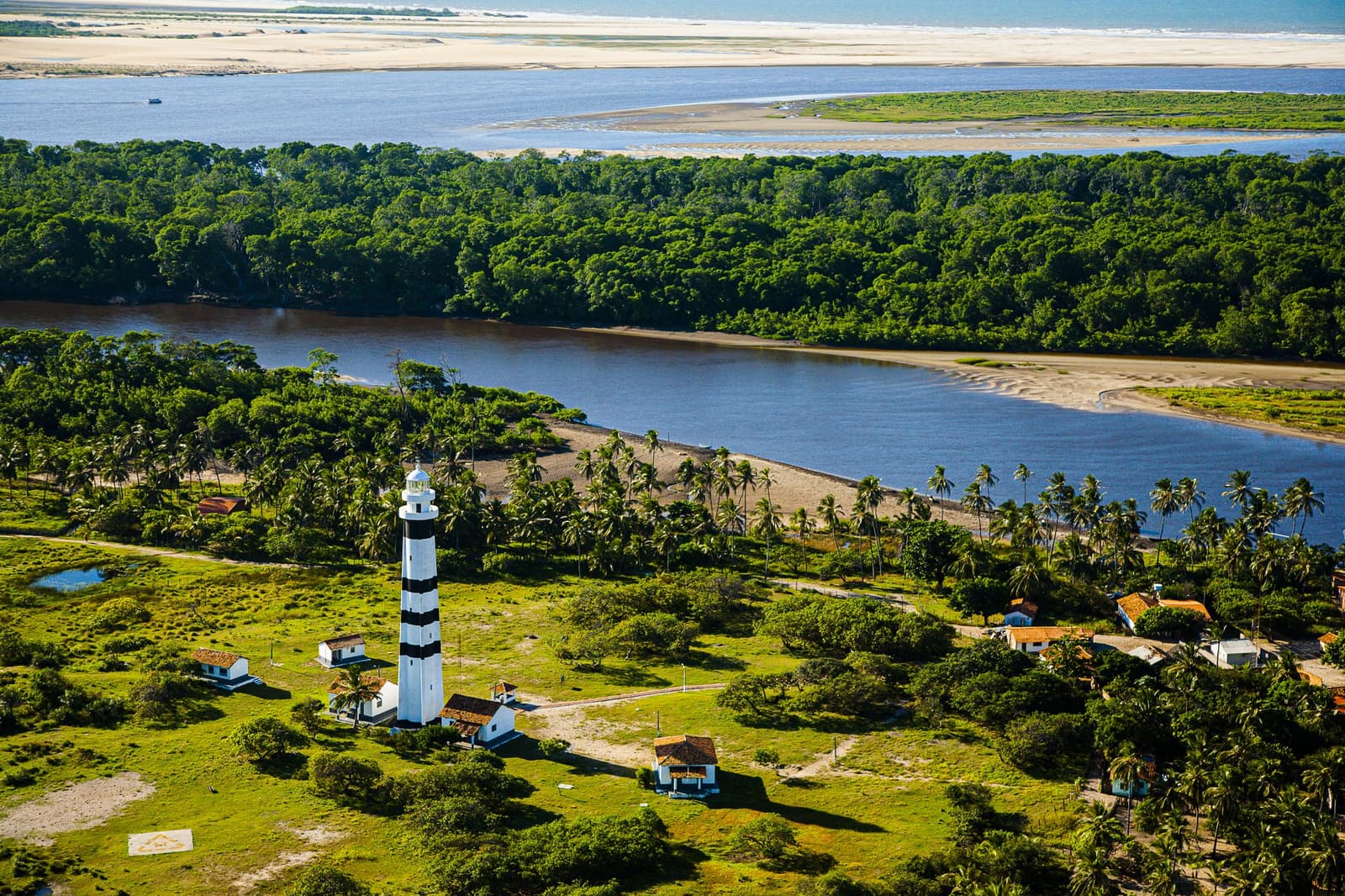 Phare rayé, cocotiers et dunes blanches à perte de vue. Photo: Gabriel Boieras Vue aérienne d'un phare rayé noir et blanc entouré de palmiers et de végétation dense, avec un large estuaire et des dunes de sable blanc s'étendant à l'horizon — repère emblématique de la randonnée dans les Lençóis Maranhenses