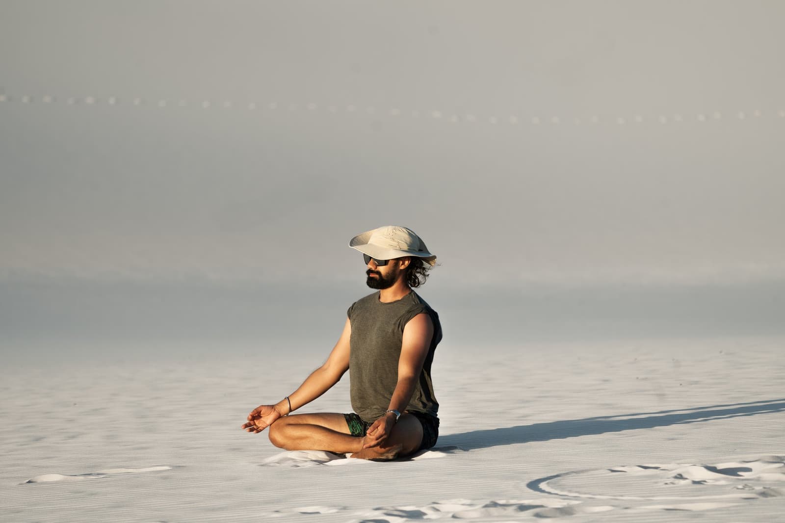 Immobile sur le sable blanc, perdu dans la vastitude du parc. Un homme barbu en casquette et lunettes de soleil est assis en tailleur sur les vastes étendues de sable blanc lors d'une randonnée dans les Lençóis Maranhenses, cerné d'un horizon pâle et infini