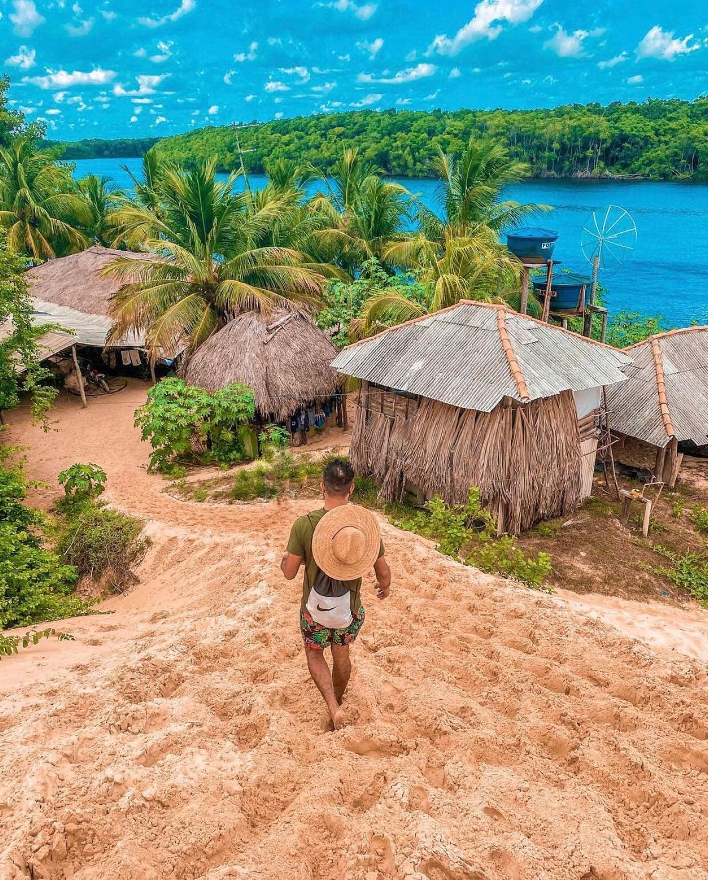 Pieds nus sur le sable chaud, un village de pêcheurs apparaît entre les palmiers. Un voyageur pieds nus, chapeau de paille et sac à dos, descend une pente de sable vers des cases au toit de chaume et des toits en tôle ondulée entourés de palmiers au bord d'une large rivière bleue lors d'une randonnée dans les Lençóis Maranhenses