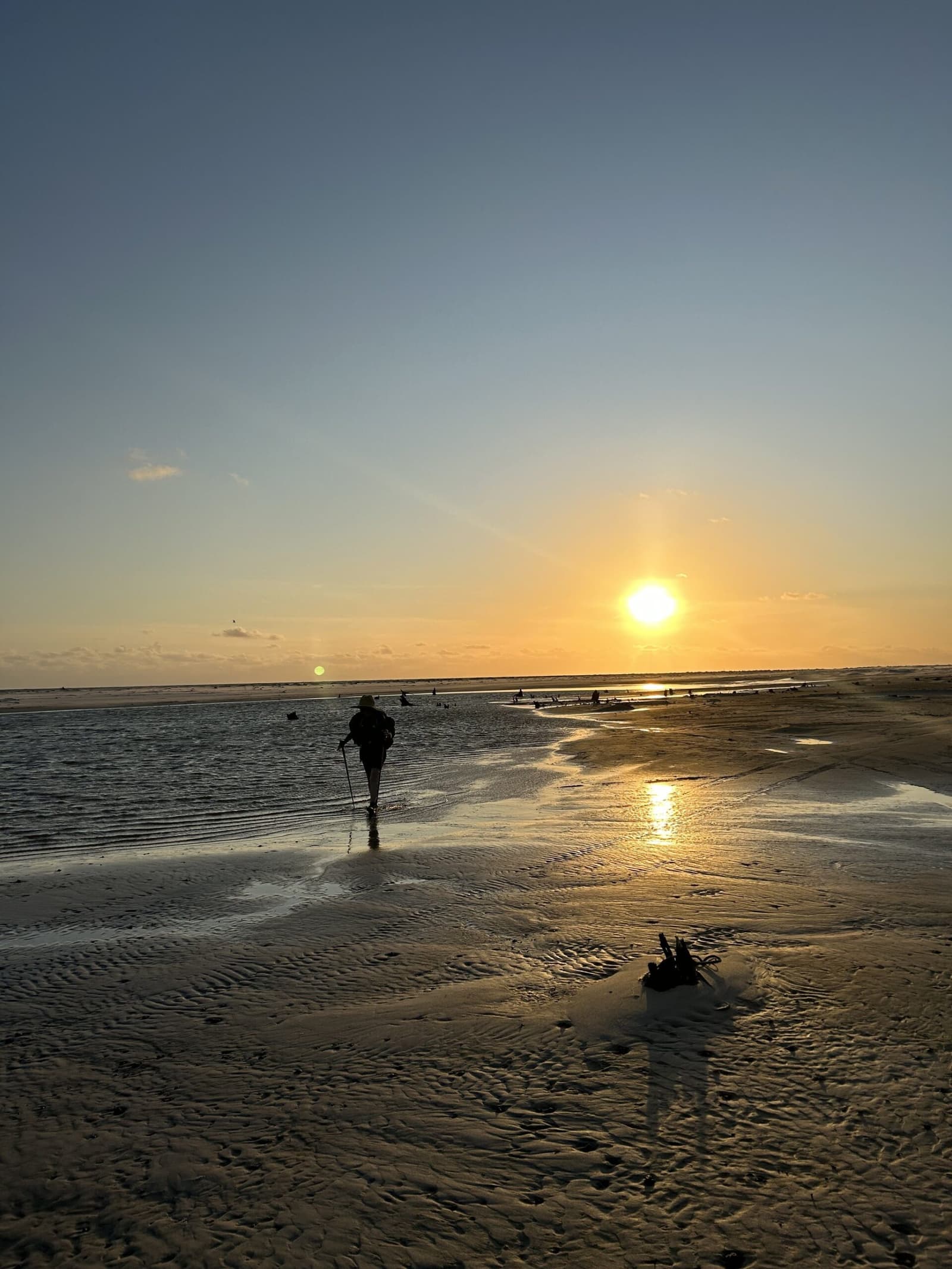 Sable mouillé, lumière dorée — un instant suspendu entre ciel et mer. Un randonneur solitaire avec sac à dos et bâtons de marche traverse à gué une eau peu profonde sur la randonnée des Lençóis Maranhenses, en silhouette contre un coucher de soleil doré sur de vastes étendues de sable ridé
