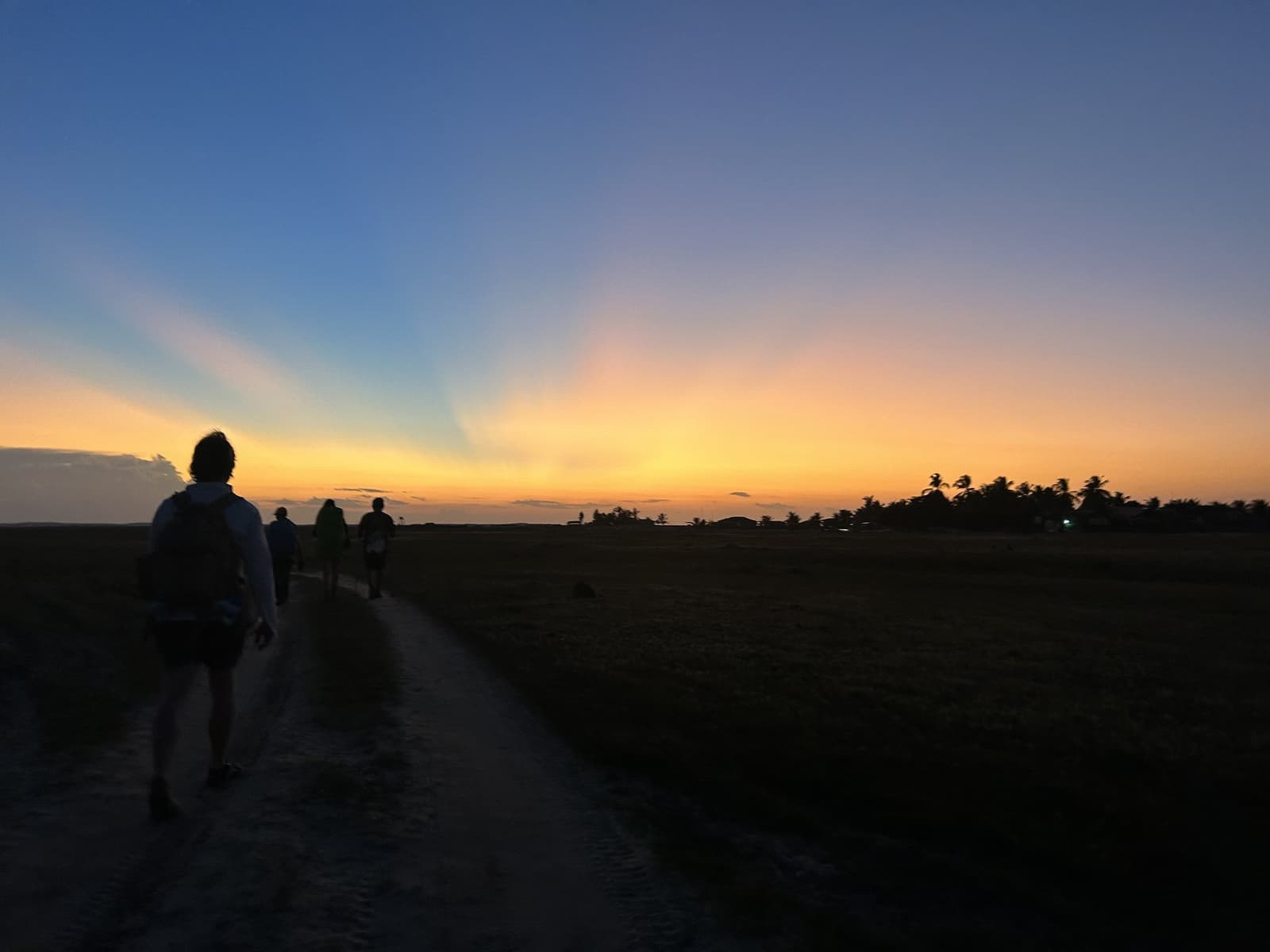 La plaine s'efface dans le crépuscule, le chemin s'allonge encore. Silhouettes de randonneurs sur un chemin de sable lors de la randonnée dans les Lençóis Maranhenses, sous un coucher de soleil aux tons orangés et bleus, avec des palmiers à l'horizon