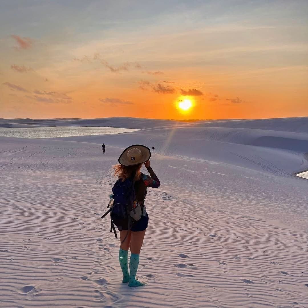 Chapeau tenu contre le vent, tout l'horizon en flammes. Une randonneuse avec sac à dos et chapeau à larges bords se tient sur les dunes blanches des Lençóis Maranhenses, face à un coucher de soleil orange vif, avec une lagune d'eau douce qui scintille à l'arrière-plan