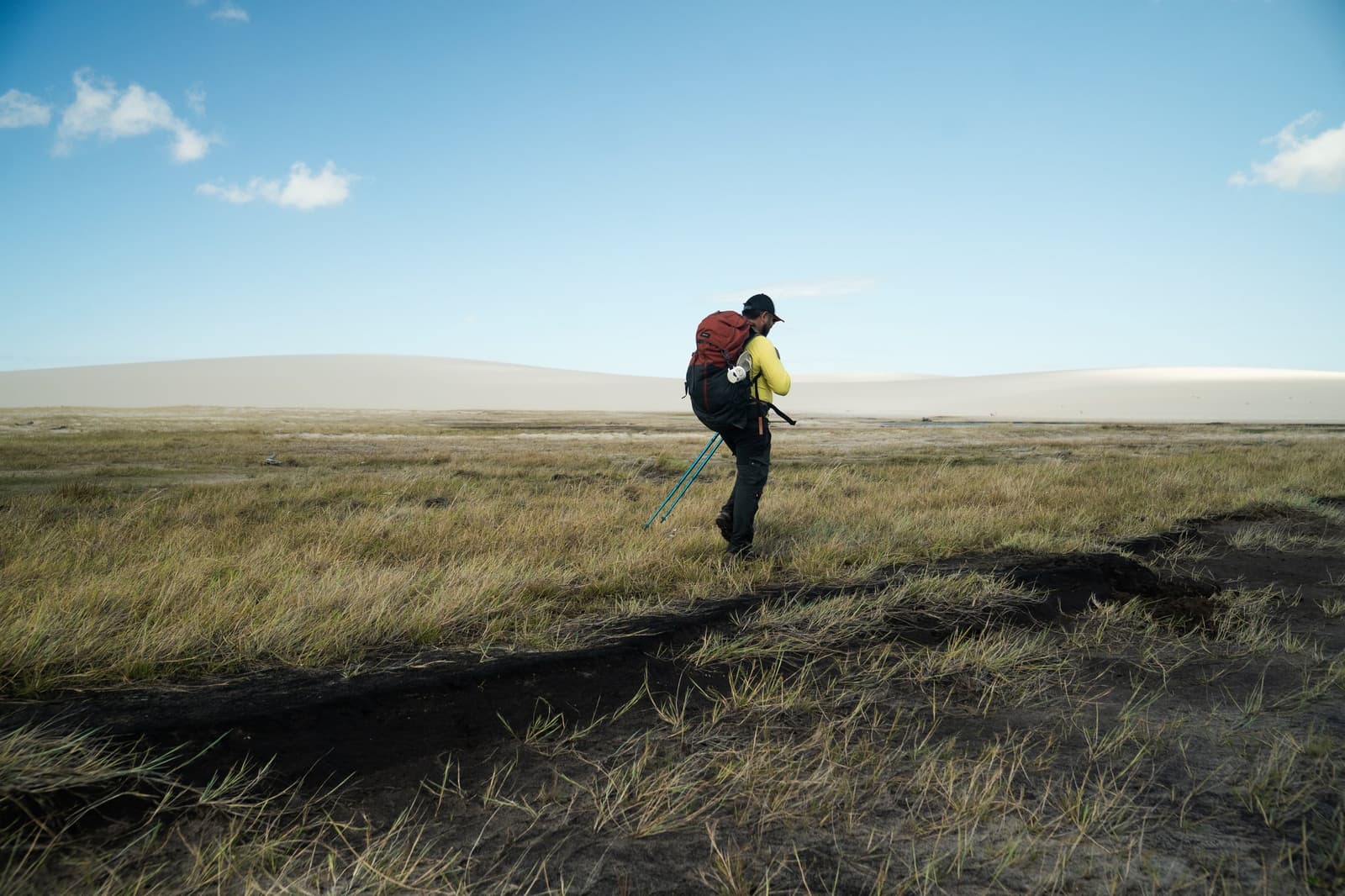 Bâtons en main, sac bien chargé — les dunes attendent au loin. Un randonneur équipé d'un grand sac à dos rouge et de bâtons de trek traverse une vaste plaine herbeuse lors de la randonnée des Lençóis Maranhenses, avec des dunes de sable pâle s'étirant à l'horizon sous un ciel bleu