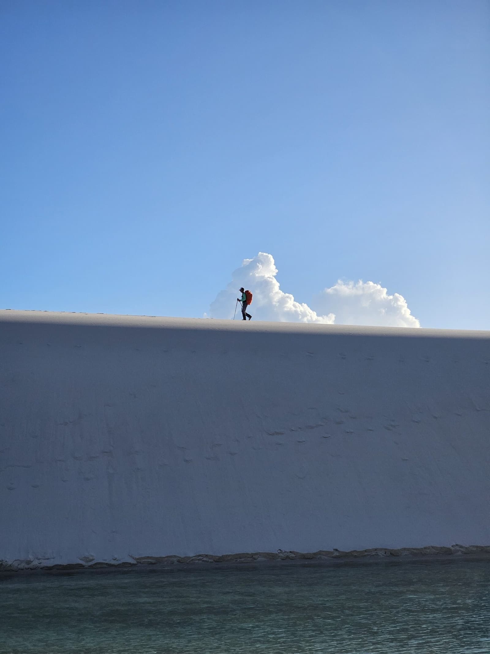 Seul sur la crête blanche, entre ciel, sable et eau. Un randonneur solitaire avec un sac rouge et un bâton de marche en silhouette sur la crête tranchante d'une dune de sable blanc éclatant lors de la randonnée dans les Lençóis Maranhenses, avec l'eau turquoise d'une lagune en bas et un cumulus imposant en arrière-plan