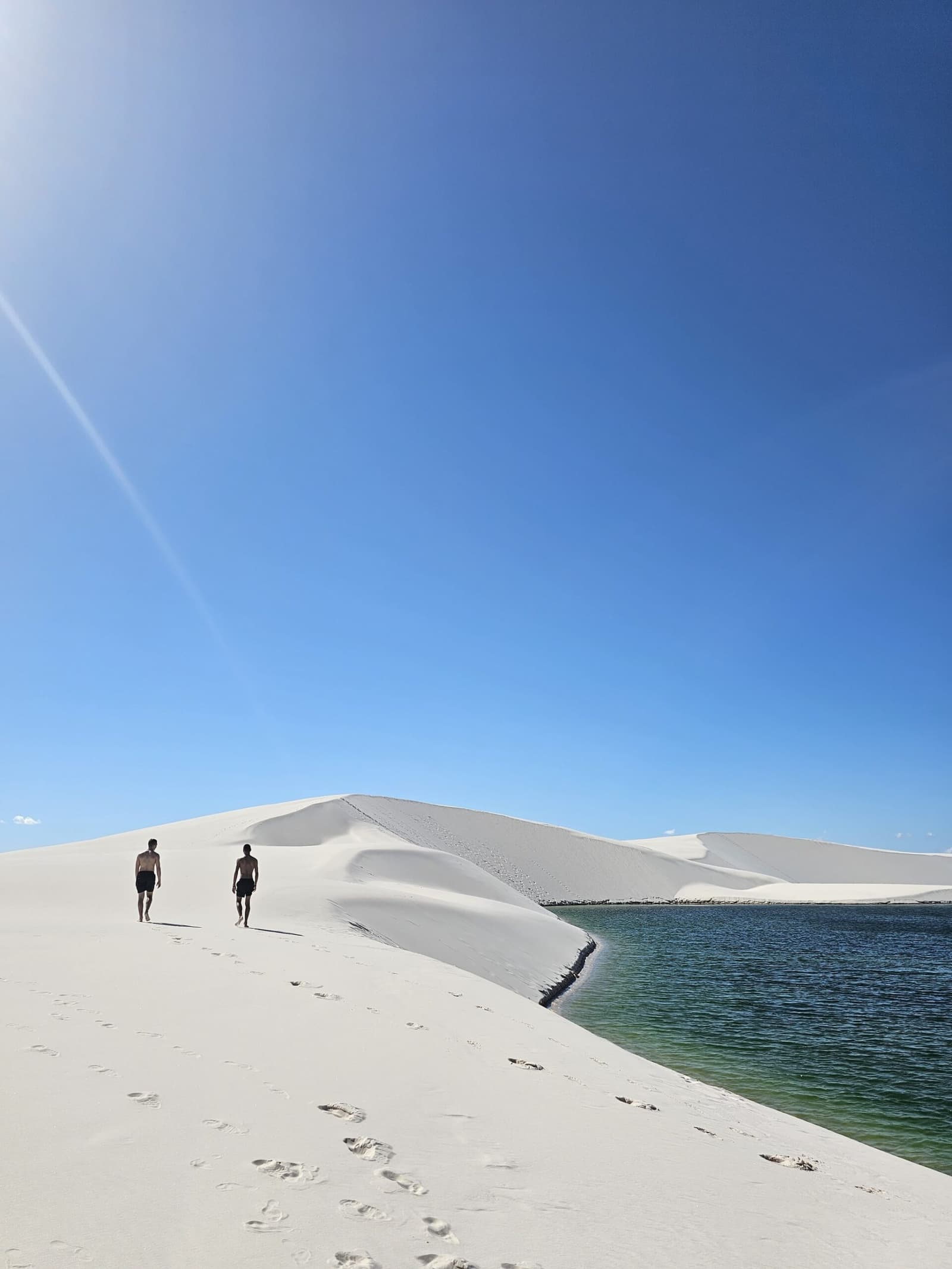 Des empreintes dans le sable blanc, une lagune émeraude à portée de main. Deux randonneurs marchant pieds nus sur des dunes d'un blanc éclatant longeant une lagune d'eau douce turquoise-verte sous un ciel bleu profond lors d'une randonnée dans les Lençóis Maranhenses