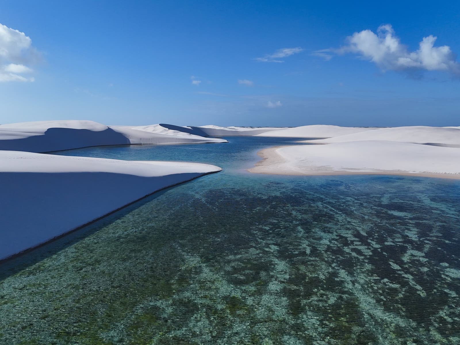 Sand and water taking turns deciding which one wins. Aerial view of a crystal-clear turquoise lagoon nestled between sweeping white sand dunes under a deep blue sky on a Lençóis Maranhenses trip