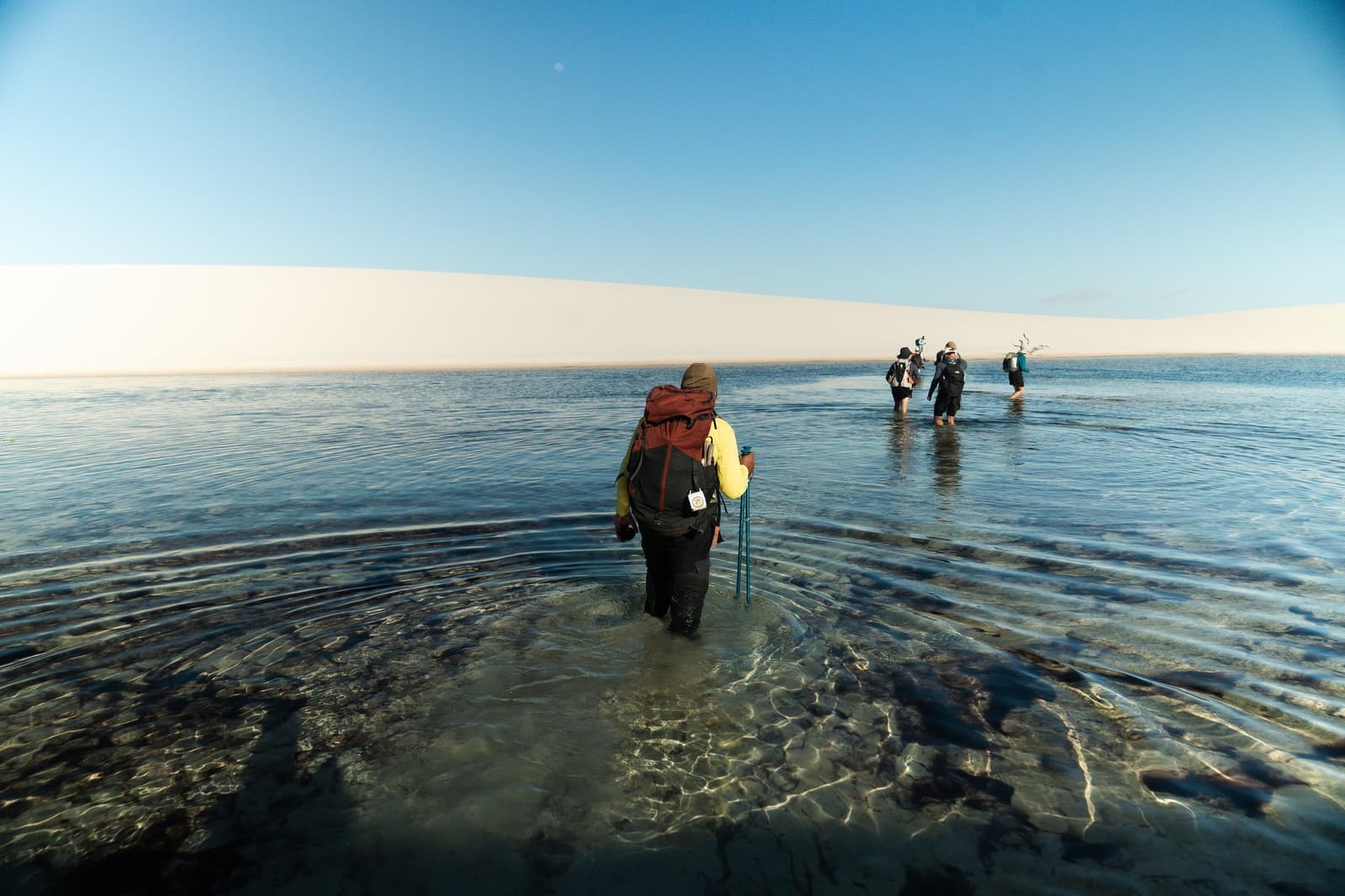 Boots off, trousers rolled, this is how you cross the dunes. A group of backpackers wading knee-deep through a clear shallow lagoon on a Lençóis Maranhenses trip, with vast white sand dunes stretching across the horizon under a blue sky