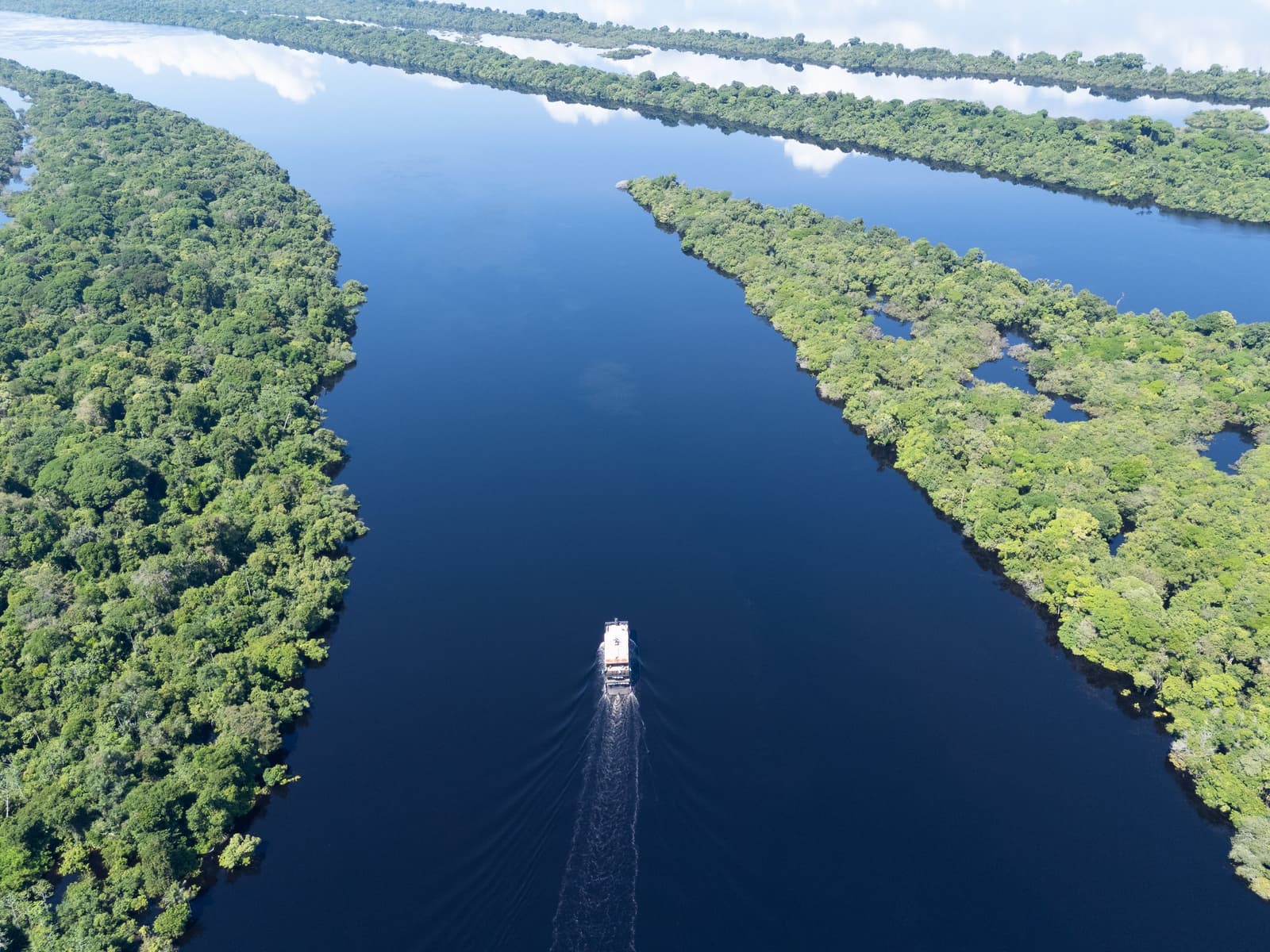 The river knows where it's going. You just get to enjoy the ride. Aerial view of a riverboat navigating a dark-water channel flanked by dense Amazon rainforest canopy on a luxury Amazon cruise in Brazil