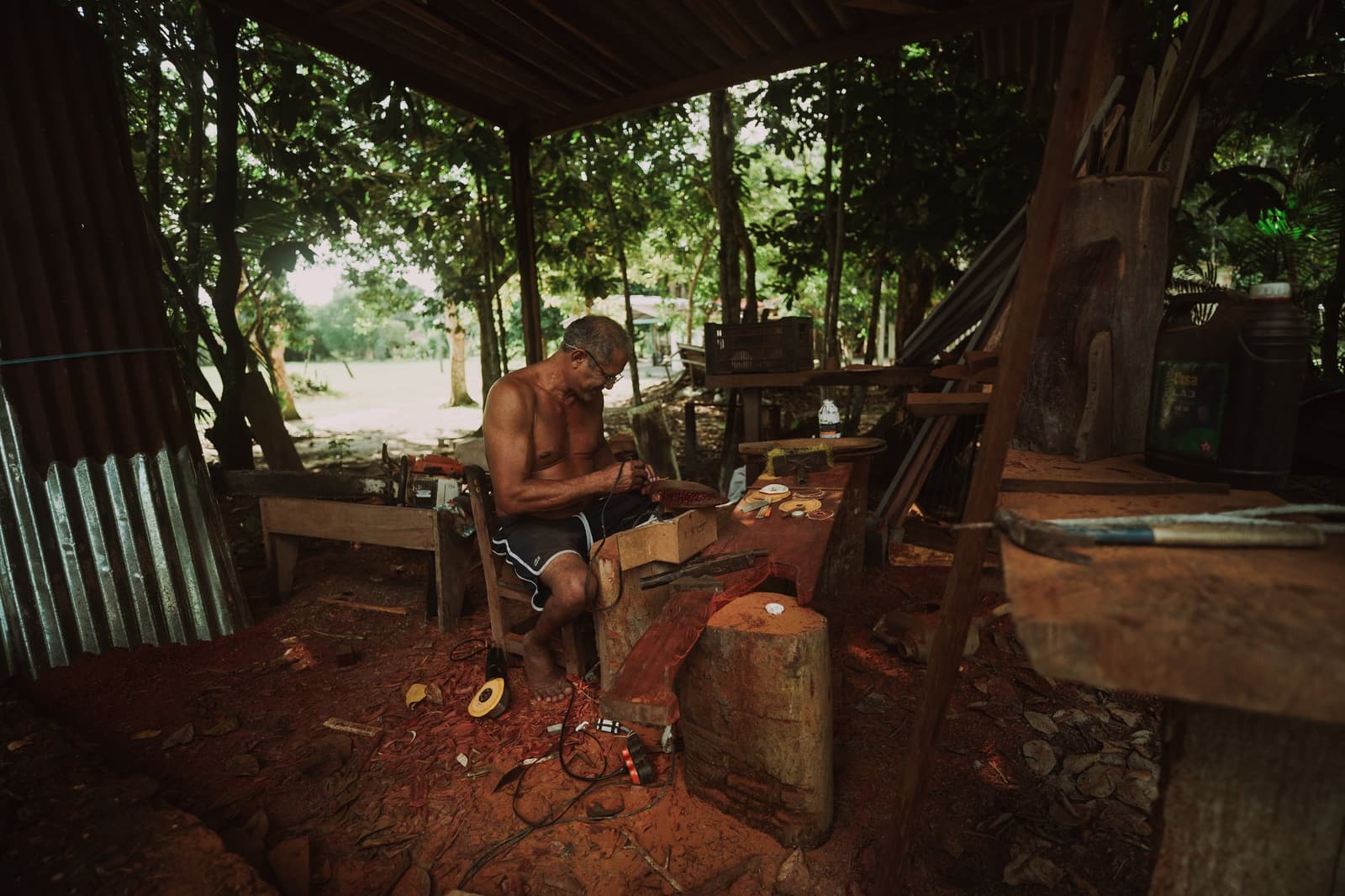 Craft passed down quietly, one careful cut at a time. A local craftsman sits shirtless in an open-air riverside workshop, carefully hand-finishing a wooden piece surrounded by tools and red-earth soil on a Luxury Amazon Cruise in Brazil community visit