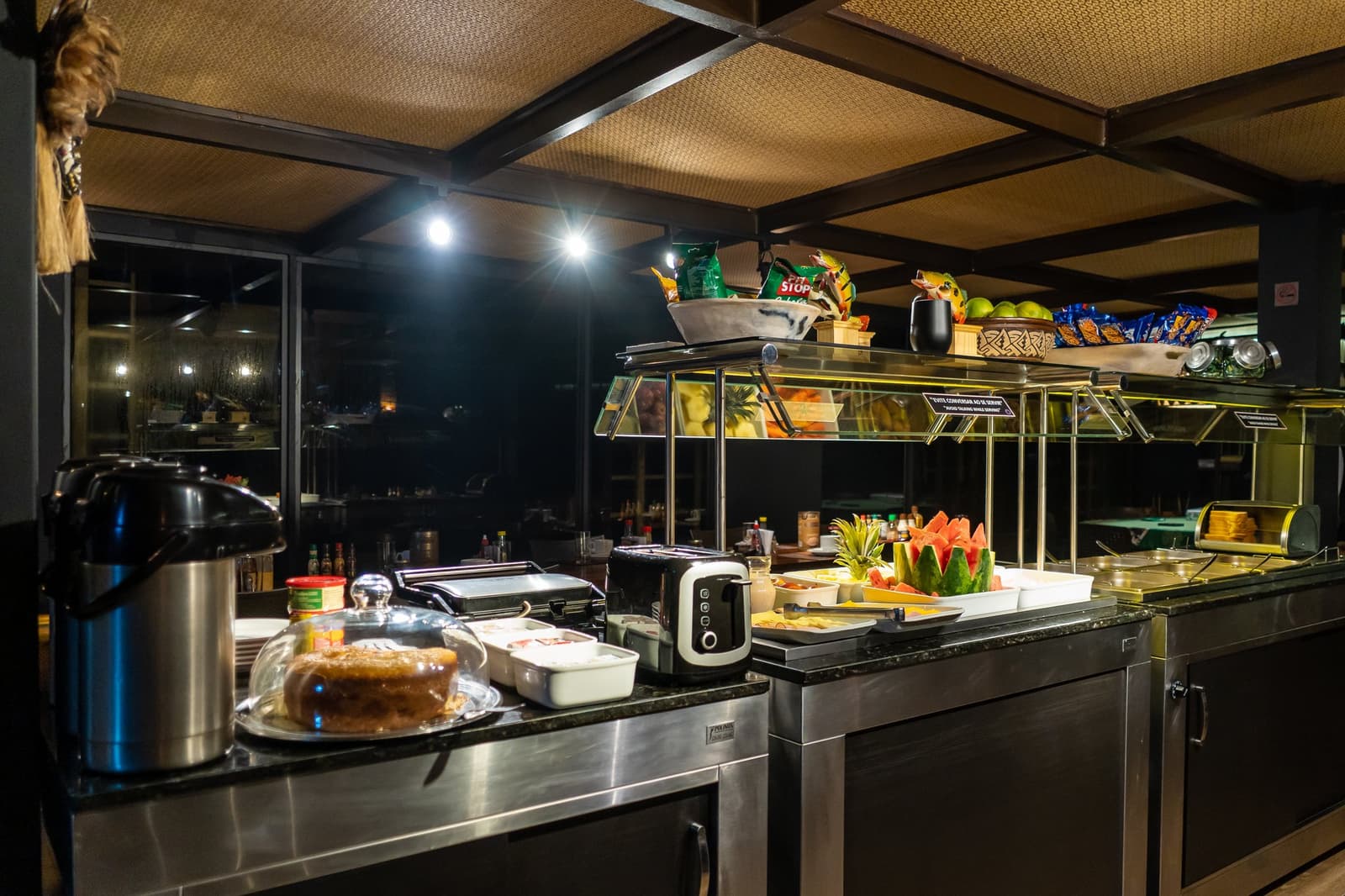Evenings on the river come with cake, fruit, and something warm in your cup. A well-lit onboard buffet on a luxury Amazon cruise in Brazil, featuring sliced watermelon, fresh tropical fruits, a whole cake under a glass dome, a toaster, thermal coffee urn, and snacks on tiered shelving, with dark river views through large windows at night