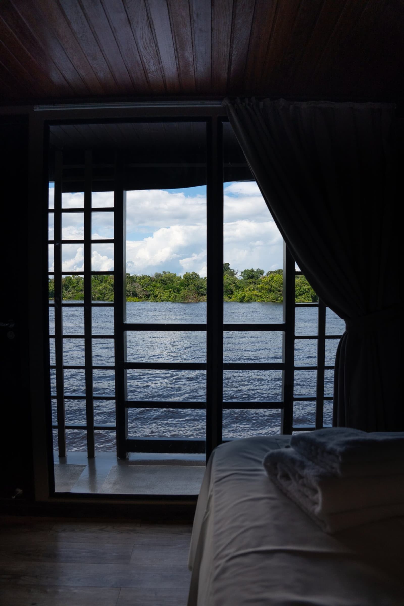 Your morning begins before you even leave the bed. View from a dark, wood-panelled suite cabin aboard a luxury Amazon cruise in Brazil, with floor-to-ceiling grid windows framing a wide river and lush green treeline under a cloudy sky
