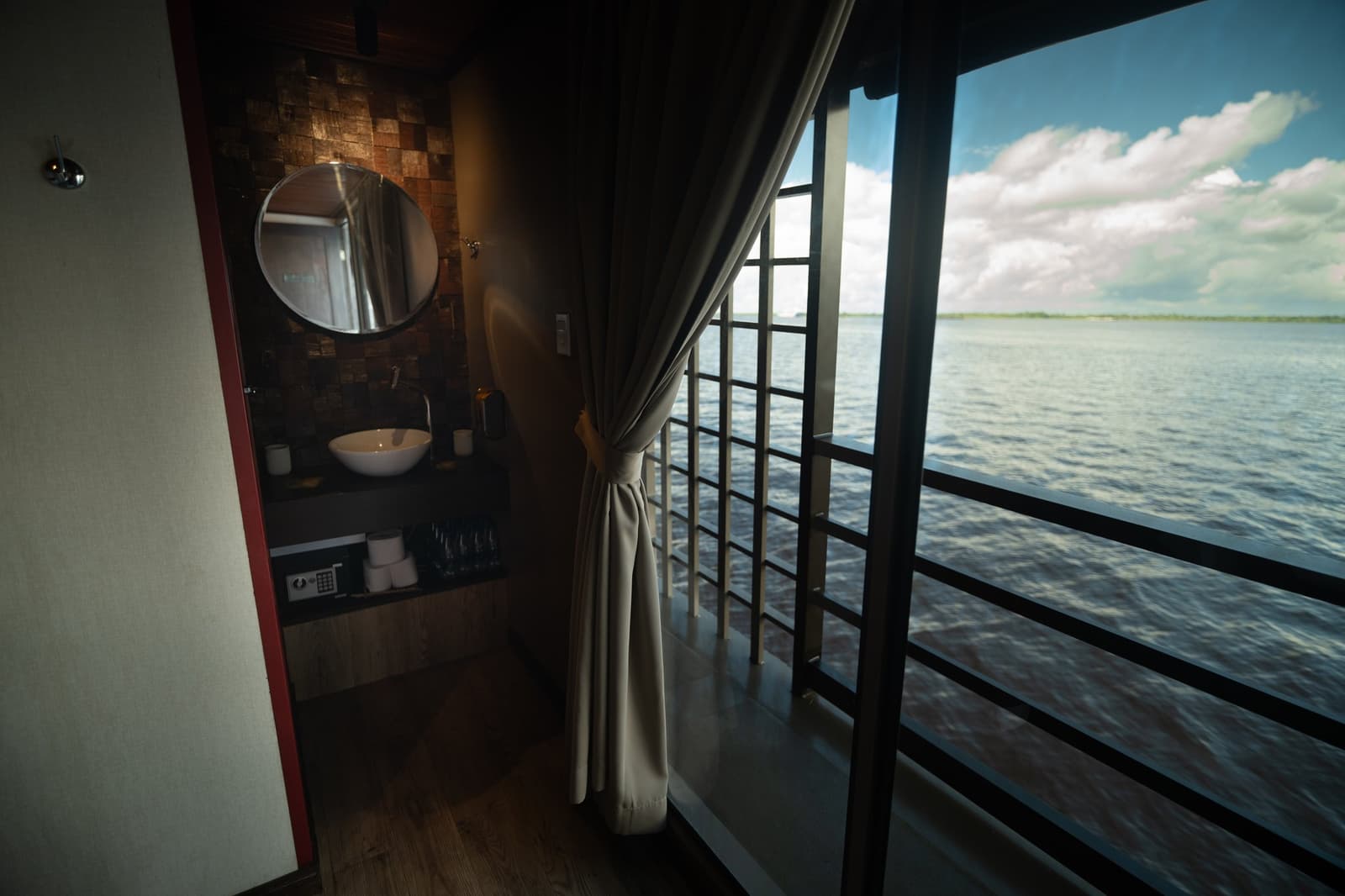 Your own slice of the river, right from the cabin door. Suite cabin interior on a luxury Amazon cruise in Brazil featuring a round mirror over a vessel-sink vanity, room safe, and floor-to-ceiling windows opening onto the wide river under a blue sky