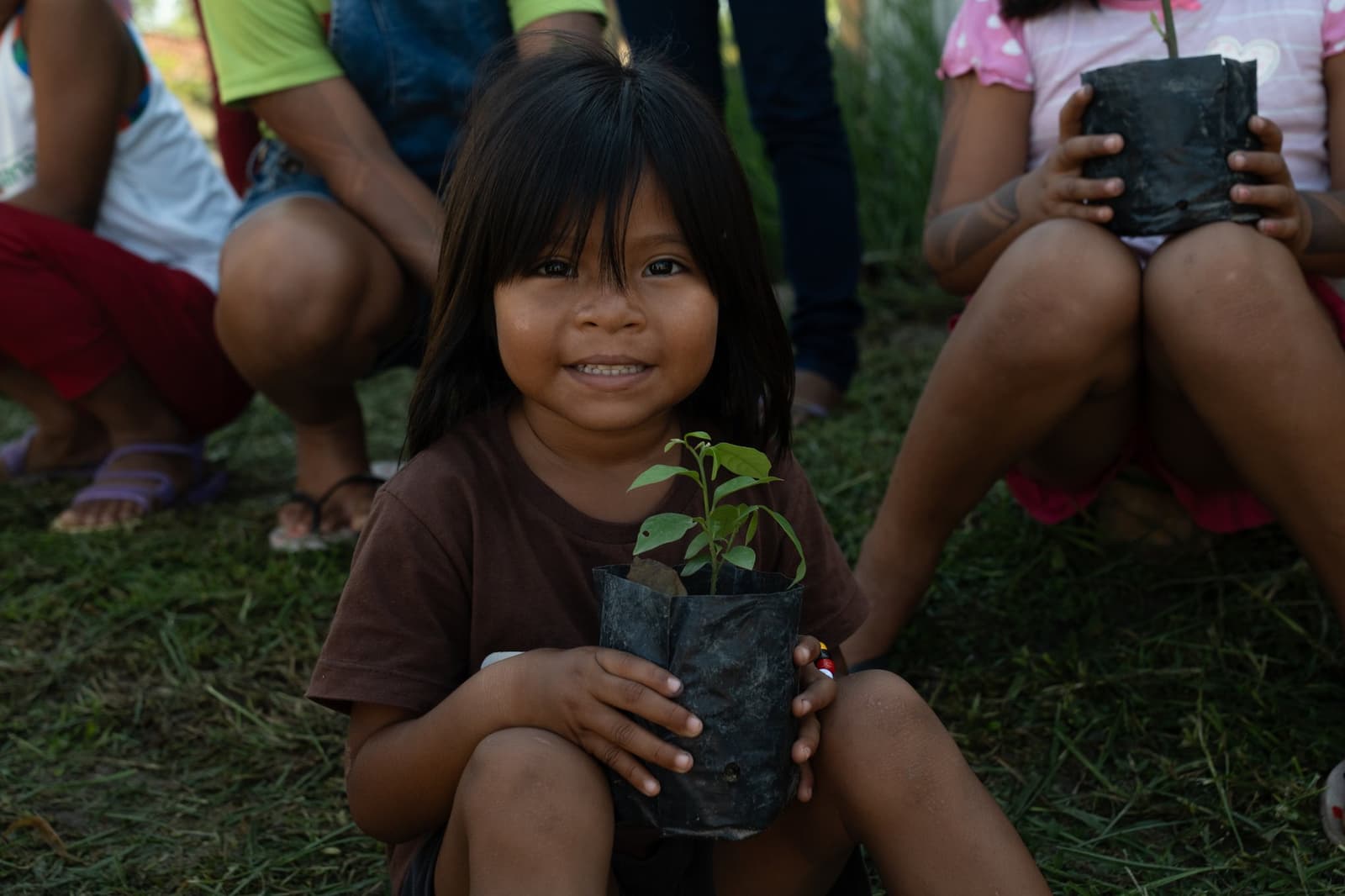 She already knows this one is hers to look after. A smiling young girl holds a green seedling in a black nursery pot during a community visit on a luxury Amazon cruise in Brazil, with other children seated on the grass behind her