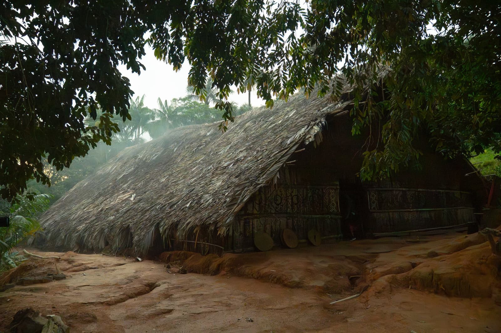 Centuries of knowledge about what makes a good roof, right here. A large palm-thatched indigenous communal longhouse with geometric painted decorations on its wooden walls, surrounded by tropical trees and red-earth ground on a Luxury Amazon Cruise in Brazil