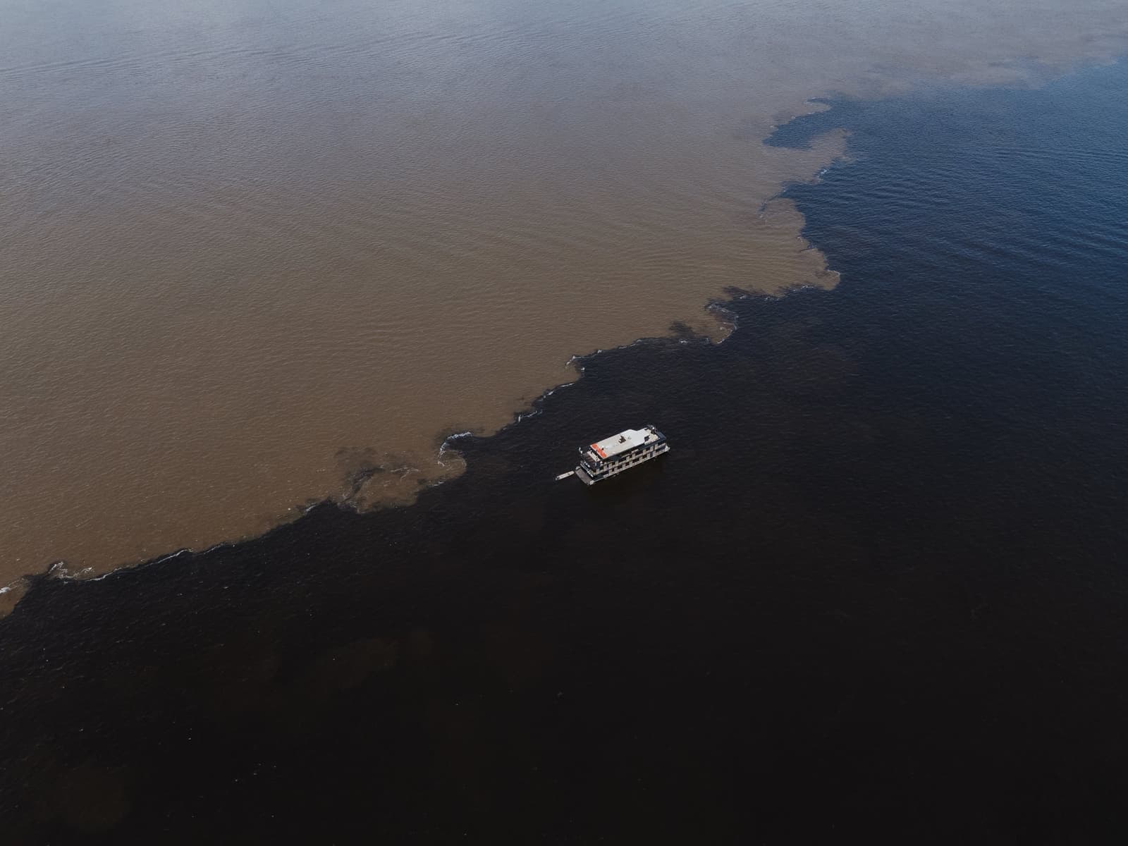 Two rivers, one boat, and a boundary that refuses to blur. Aerial view of a riverboat crossing the Meeting of Waters on a luxury Amazon cruise in Brazil, where dark black-water and sandy brown-water rivers meet in a sharp, winding boundary across the frame