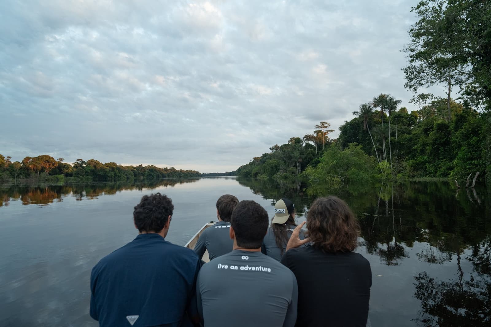 The forest answers every question before you think to ask it. Four travellers seated at the bow of a small boat on a calm, dark-water river, surrounded by dense rainforest canopy on both sides during an Amazon river cruise in Brazil