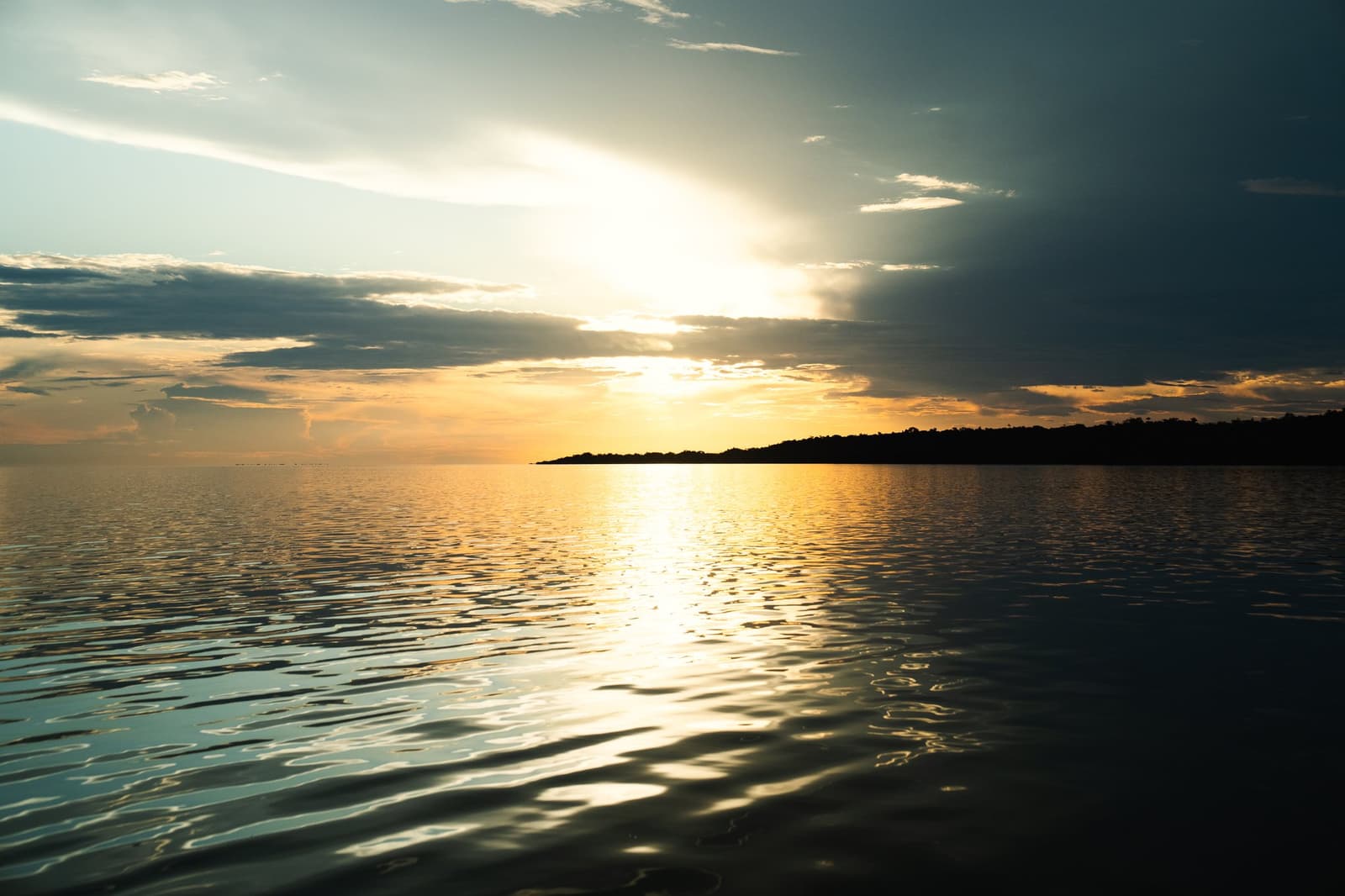 The kind of evening that makes you put your phone down and just watch. Golden sunset reflected across a vast, calm river surface on a luxury Amazon cruise in Brazil, with a dark forested island silhouetted on the horizon beneath dramatic clouds