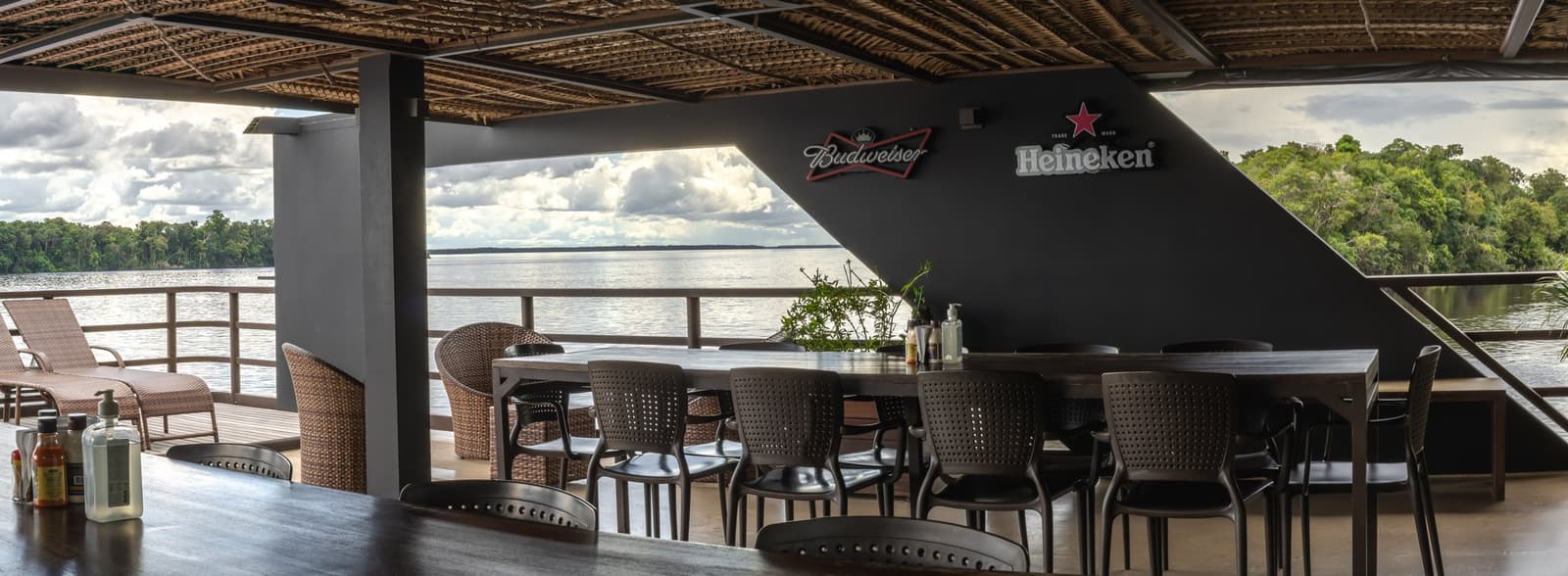 Cold drink in hand, forest on the horizon, the river does the rest. Open-air bar deck on a luxury Amazon cruise in Brazil, with rattan chairs, long dining tables under a woven-palm ceiling, and a wide river framed by dense jungle on both sides