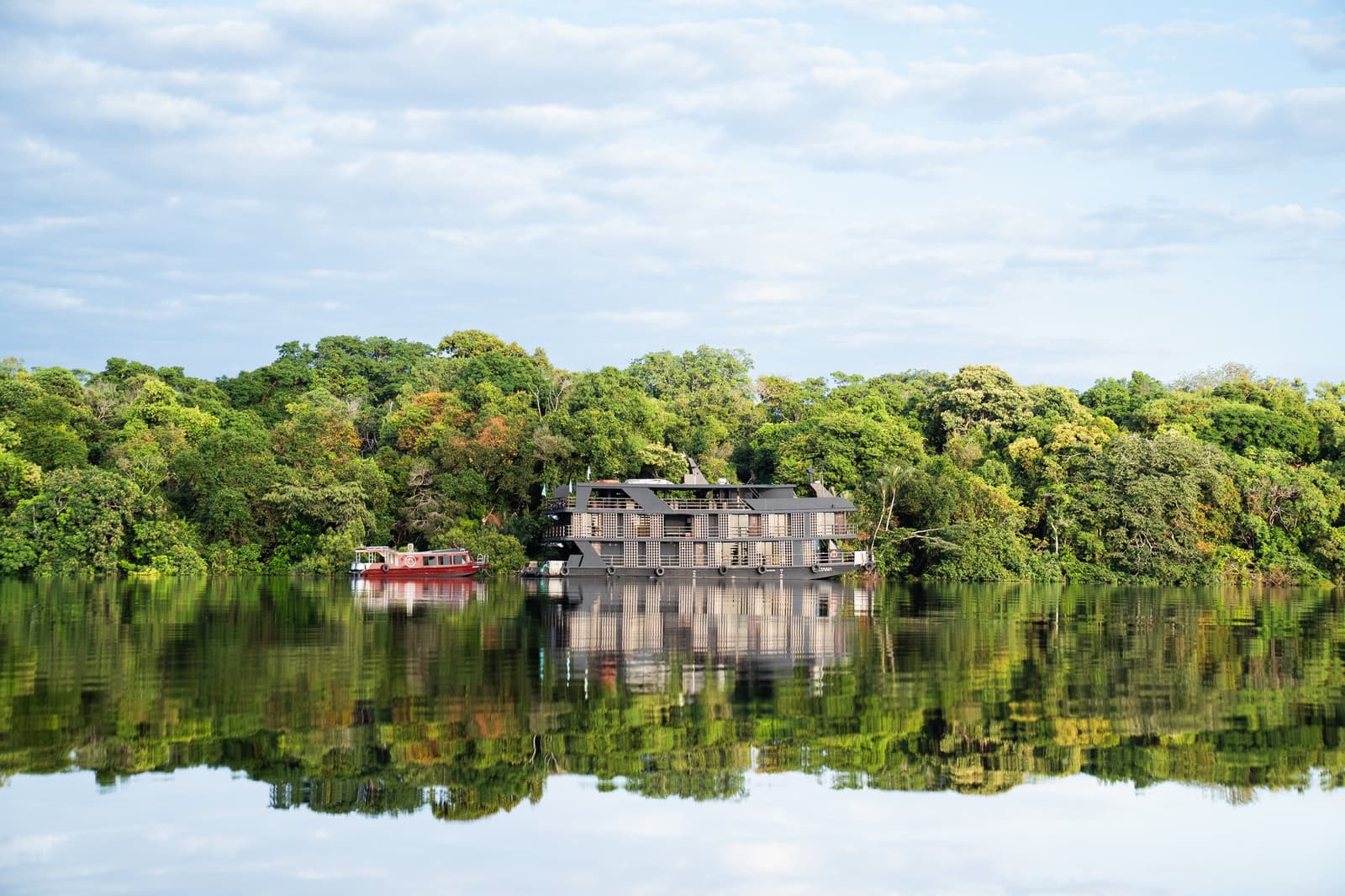 Your floating base camp, hidden in plain sight on the river. A multi-deck luxury Amazon cruise riverboat moored along a lush tropical forest riverbank, with perfect mirror reflections on the calm dark water and a red support boat alongside