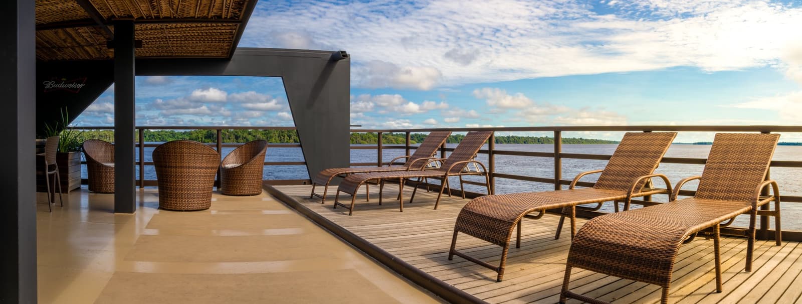 Pick a lounger, watch the Amazon slide past. Simple as that. Wicker sun loungers and rattan chairs arranged on the upper sun deck of a luxury Amazon cruise in Brazil, overlooking a wide dark-water river lined with dense rainforest under a blue sky