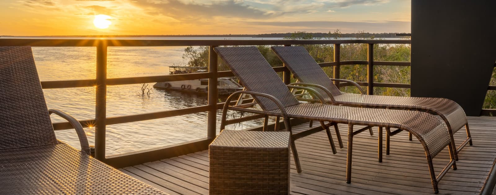 Your sundowner seat is already waiting, no reservation needed. Wicker sun loungers on a wooden deck overlooking a wide river at golden-hour sunset during a luxury Amazon cruise in Brazil