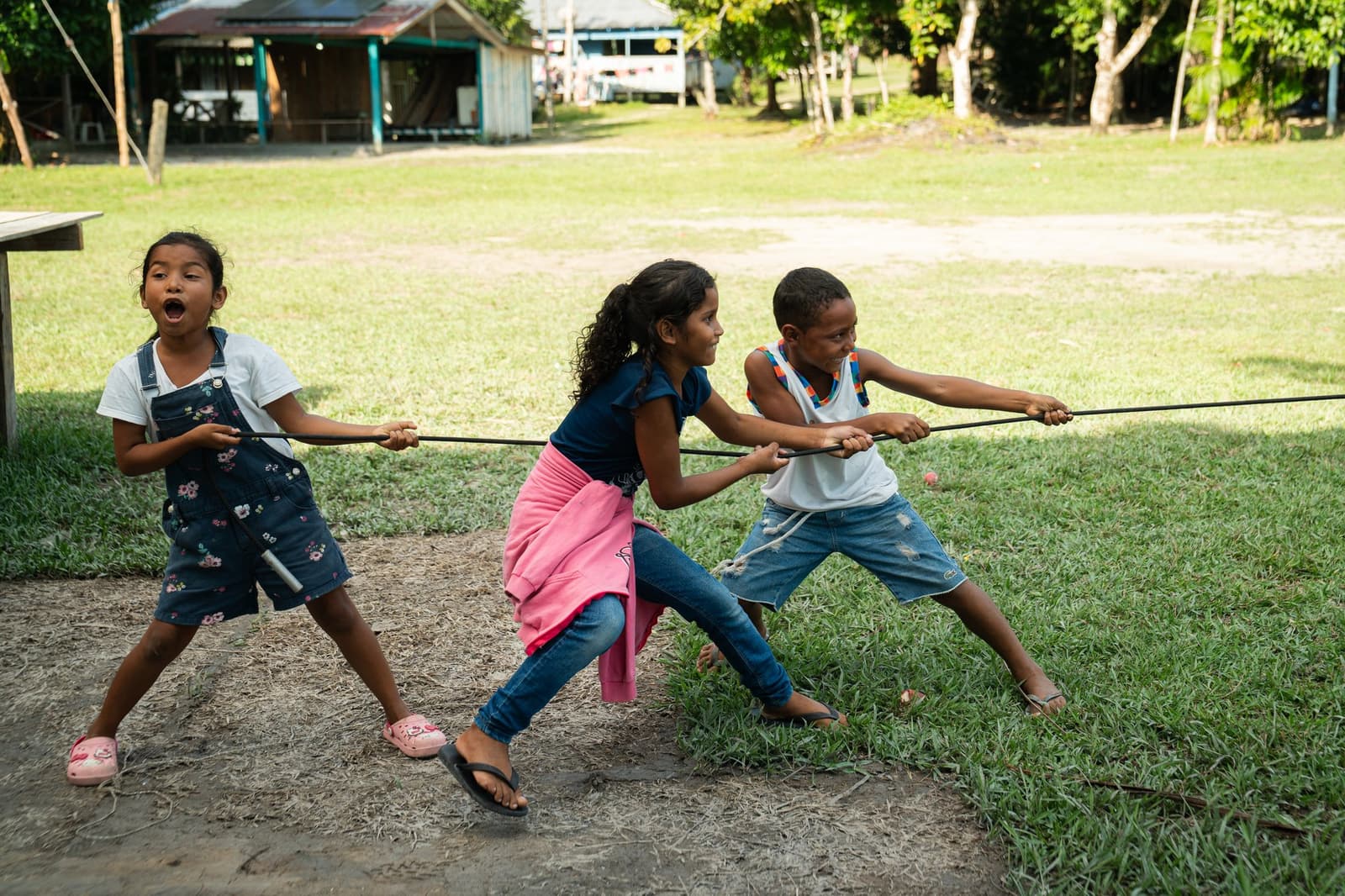 Nobody is holding back, least of all the girl on the left. Three children laughing and straining on a rope in a tug-of-war game in a riverside village clearing, a highlight of community visits on a Luxury Amazon Cruise in Brazil