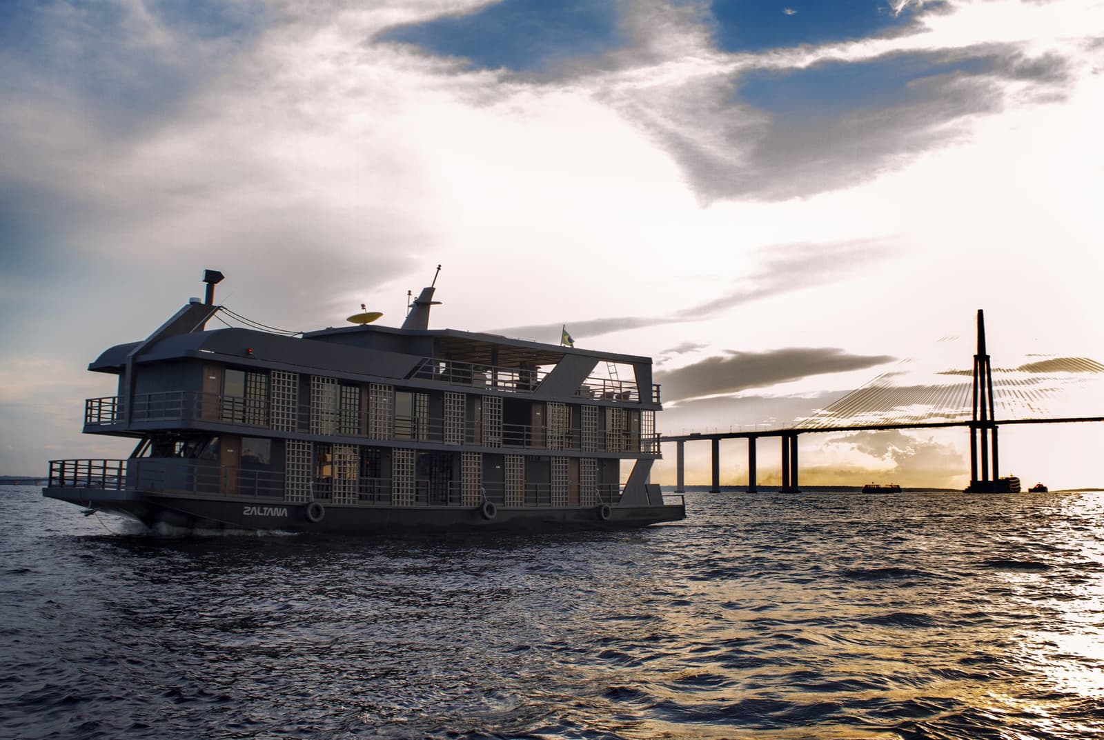 Your floating base camp, already headed somewhere worth it. The Zaltana riverboat setting out on a luxury Amazon cruise in Brazil, with a cable-stayed bridge silhouetted against a dramatic sunset sky on the broad Rio Negro
