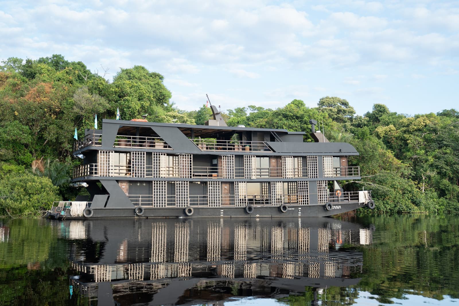 Your floating base camp, parked where the forest meets the river. The Zaltana luxury Amazon cruise riverboat moored on a glassy dark-water river, its modern multi-deck dark hull and latticed cabin balconies reflected perfectly in the still water, backed by dense rainforest canopy under a partly cloudy sky
