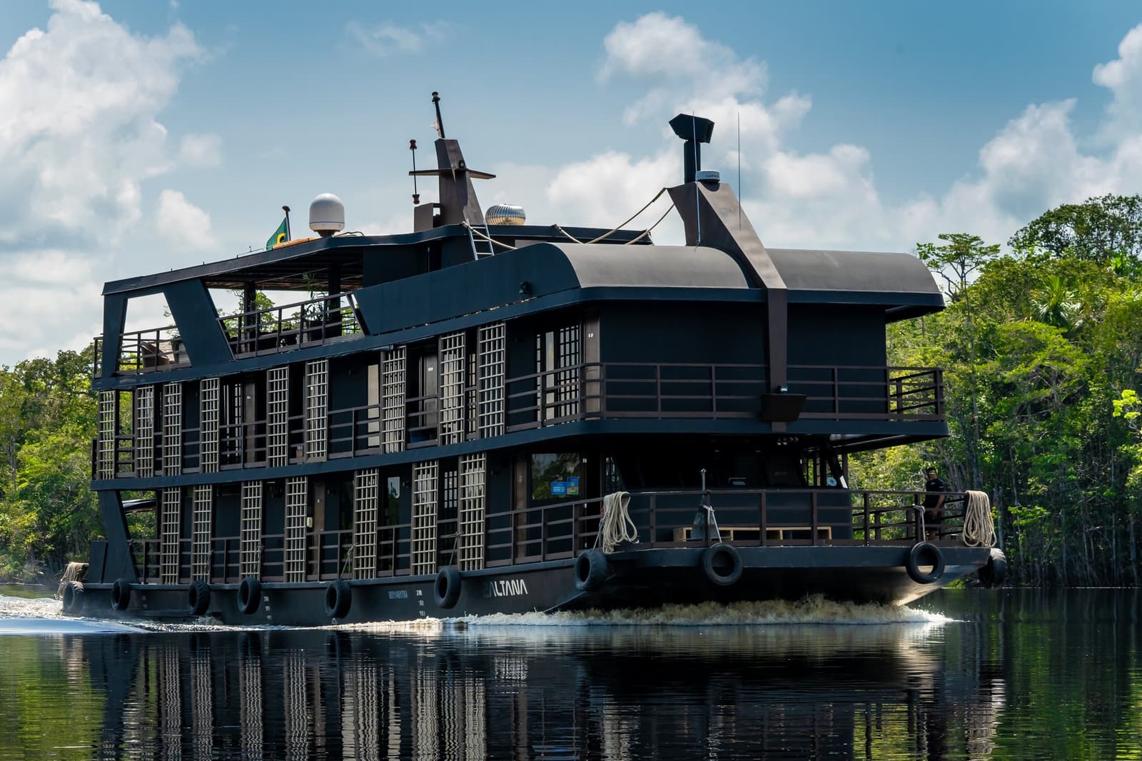 Your floating base camp, surprisingly well-stocked with comfort. The Zaltana riverboat navigating a calm dark-water channel on a luxury Amazon cruise in Brazil, surrounded by dense rainforest under a partly cloudy sky
