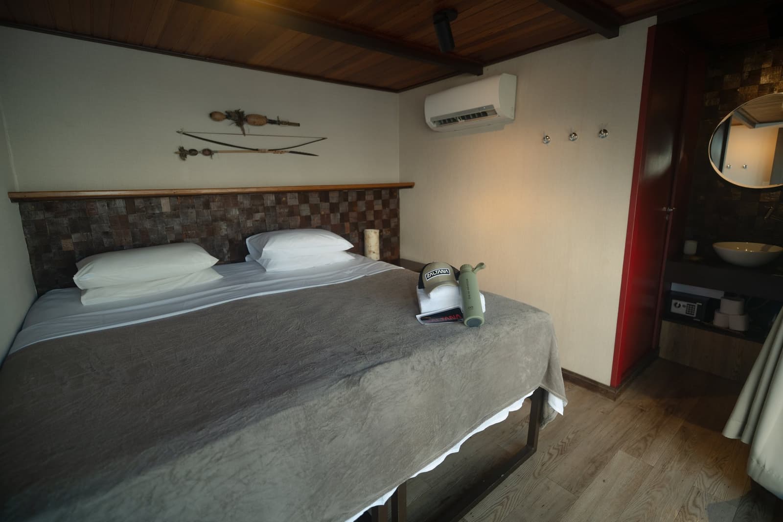 Your cabin after a long day on the river, compact, cool, and ready for you. A neatly made double bed with white pillows and grey blanket inside a Luxury Amazon Cruise in Brazil suite cabin, featuring a mosaic wood headboard, indigenous bow-and-arrow wall art, wooden ceiling, air conditioning unit, and a binoculars case resting on the bed