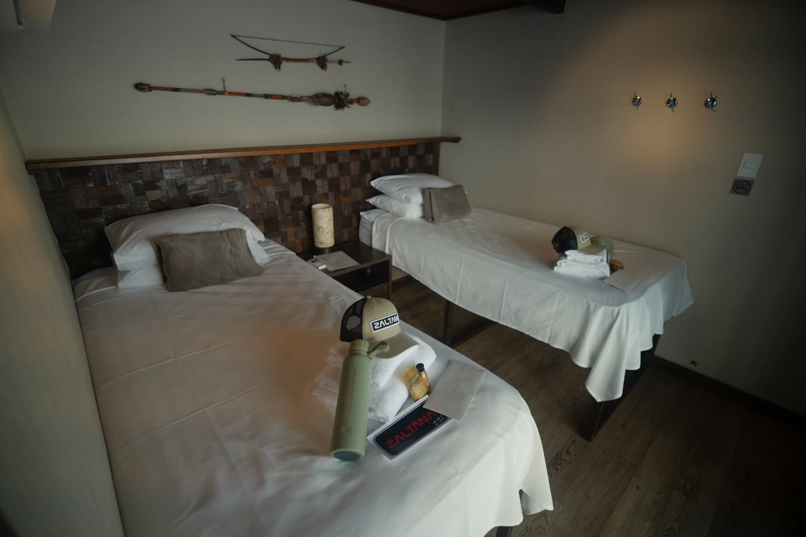 Gear dropped, beds made, the jungle can wait five more minutes. Twin-bed suite cabin aboard the Zaltana riverboat on a Luxury Amazon Cruise in Brazil, featuring crisp white linen, a stone-tile headboard, indigenous decorative artefacts on the wall, and Zaltana-branded guest amenities on the beds