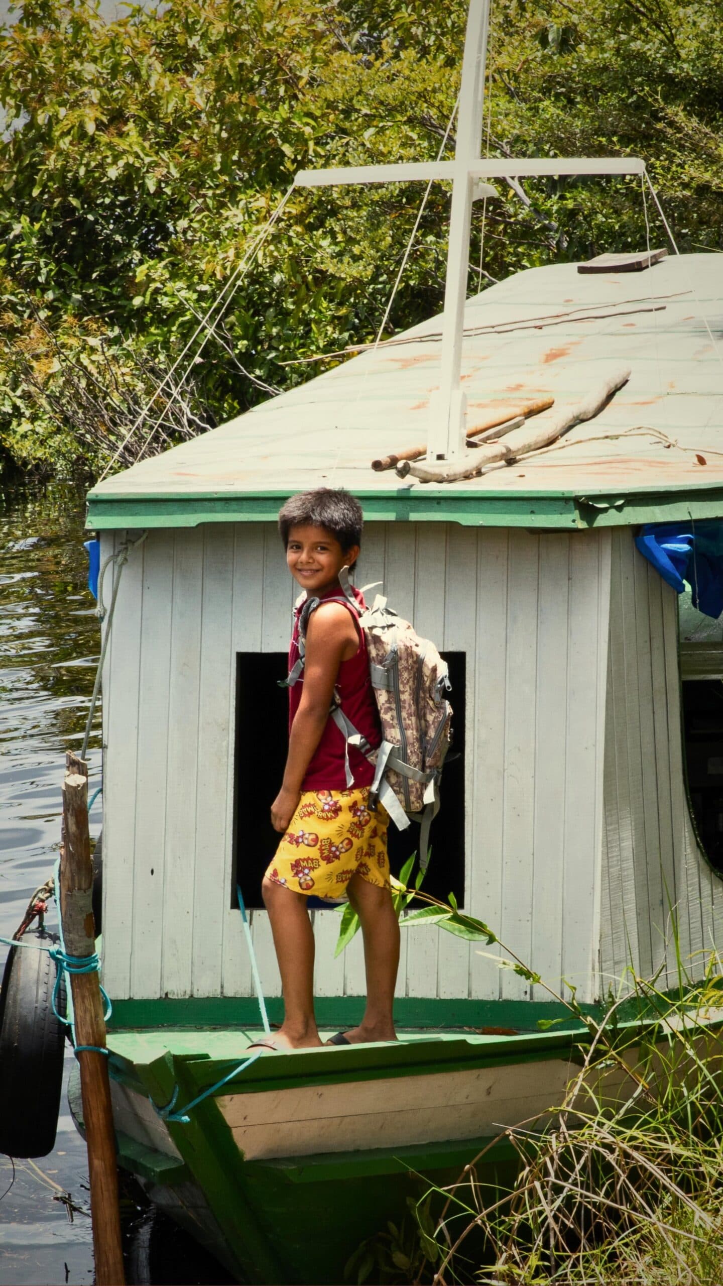 School run, Amazon style, the river is the road here. A smiling boy with a backpack stands on the deck of a small wooden riverboat moored beside lush greenery, reflecting the warm community spirit found near a luxury Amazon lodge in Brazil