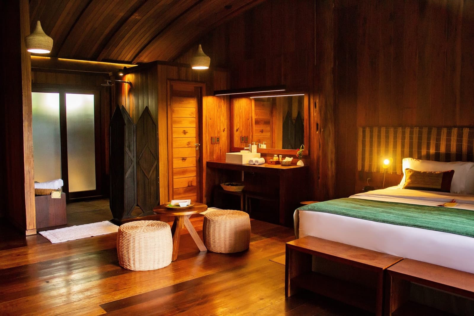 The kind of room that makes you forget you packed an alarm clock. Warmly lit timber cabin interior at a luxury amazon lodge brazil, featuring a queen bed with green throw, wicker ottomans, open vanity with white basin, and polished hardwood floors beneath an arched wooden ceiling