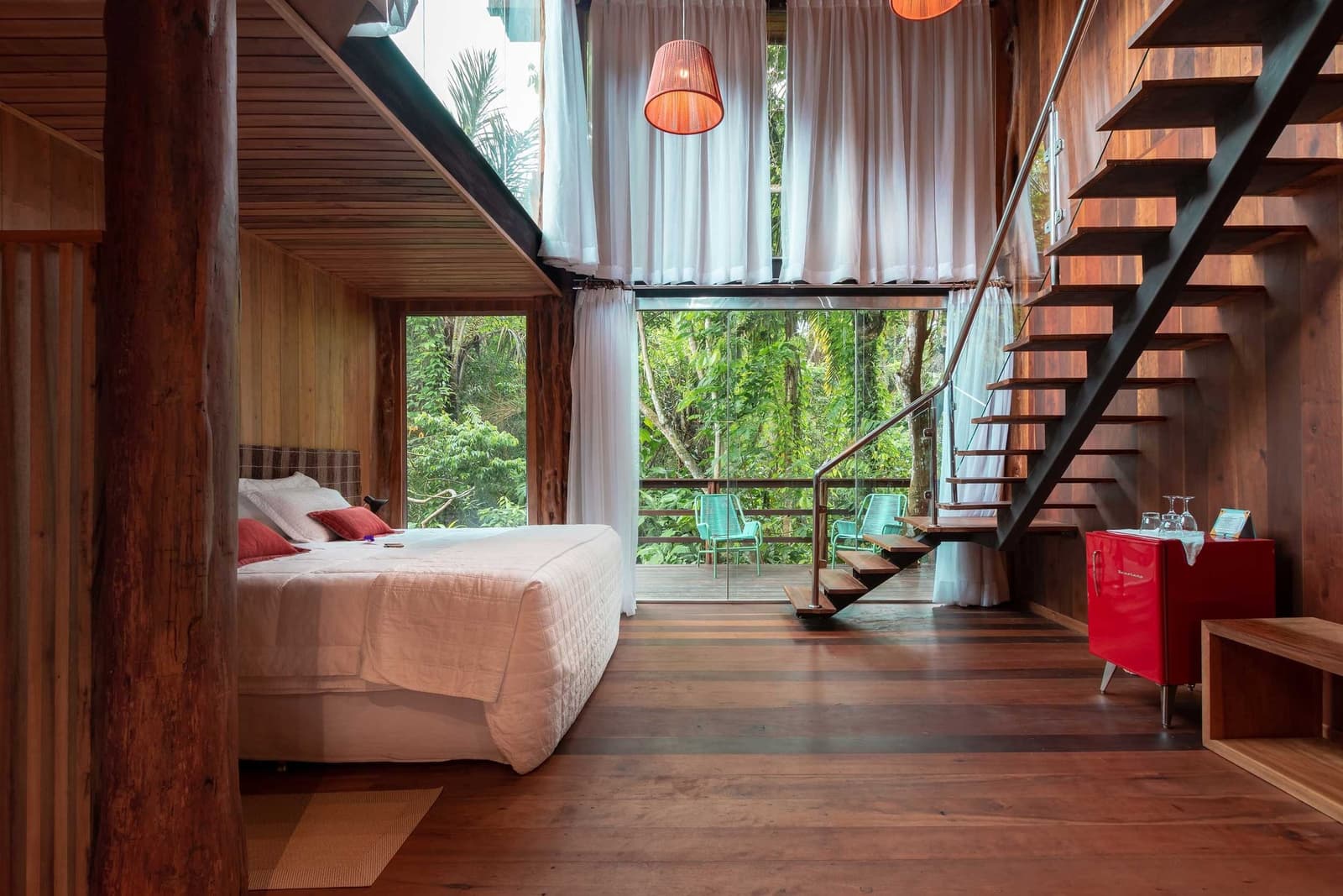 Your morning starts with a canopy view from bed, the forest does the rest. Mezzanine loft bedroom at a luxury Amazon lodge in Brazil, featuring warm hardwood walls and floors, a queen-size bed with red accent cushions, sheer white curtains, a floating staircase, a retro red minibar, and floor-to-ceiling glass doors opening onto a timber deck surrounded by dense rainforest canopy