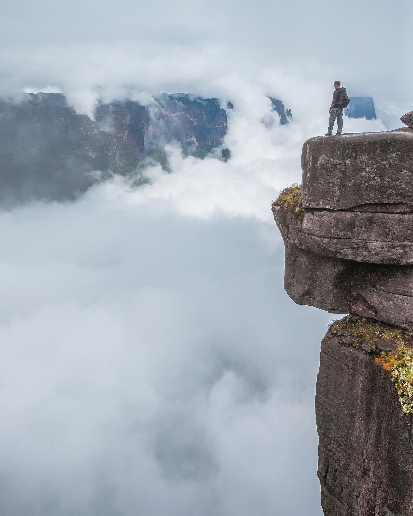 Up here, the clouds are below you and the world feels very small. A lone hiker with a backpack stands on the edge of an ancient sandstone rock pillar high above a sea of clouds on a Mount Roraima hike, with a massive tepui escarpment emerging from the mist in the distance