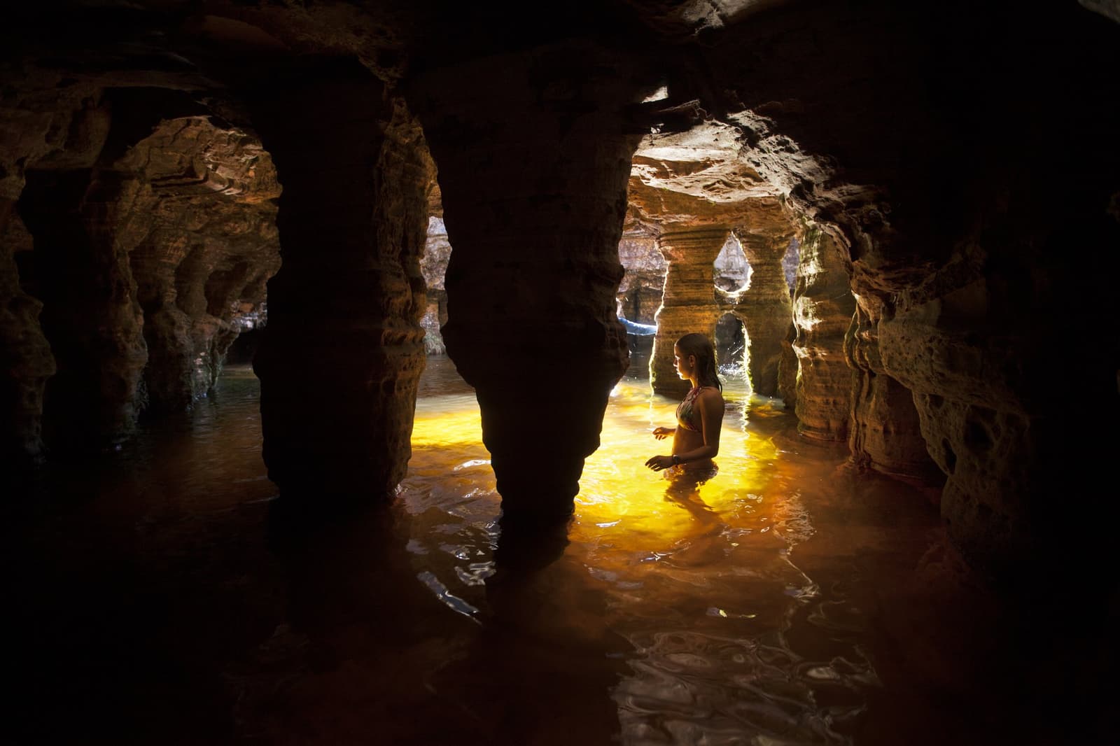 The cave keeps its own clock, and the water is warmer than you'd expect. A woman wading waist-deep through a glowing amber pool inside an ancient sandstone cave chamber, lit by warm golden light filtering through eroded rock arches on the Mount Roraima hike