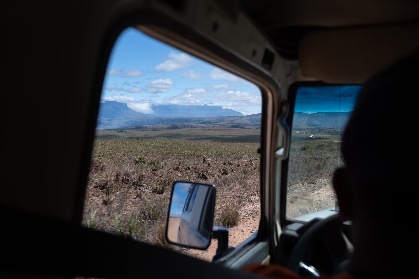 Le tepui pointe à l'horizon, et la piste, elle, ne se presse pas. Vue depuis la fenêtre d'un 4x4 sur une immense savane ouverte avec un tepui à sommet plat à l'horizon lors de l'approche du trek du Mont Roraima