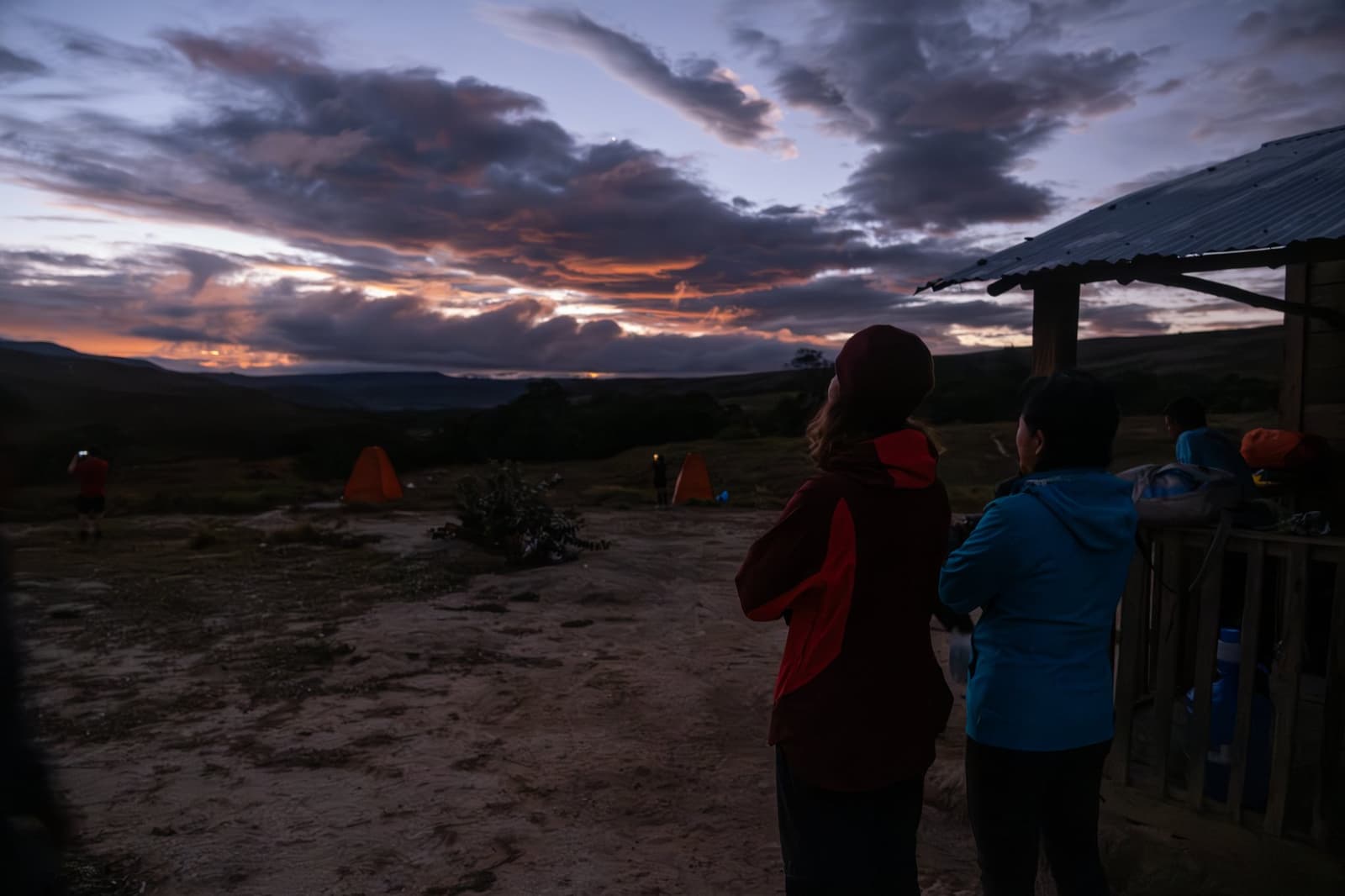 Les jambes fatiguées, les yeux grands ouverts sur ce ciel du soir. Des randonneurs en vestes chaudes observent un coucher de soleil spectaculaire orange et violet depuis un campement en flanc de colline lors d'un trek au Mont Roraima, avec des tentes orange lumineuses en arrière-plan