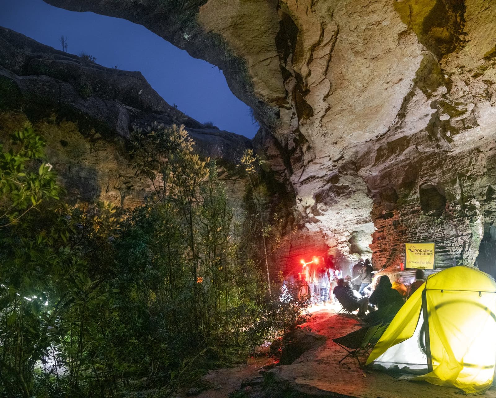 Un abri de grès vieux de 70 millions d'années, et une nuit comme aucune autre. Des randonneurs réunis autour d'un feu de camp à l'intérieur d'un immense abri rocheux en grès la nuit lors d'un trek au mont Roraima, avec une tente jaune lumineuse au premier plan et une bannière Roraima Adventures visible sur la paroi