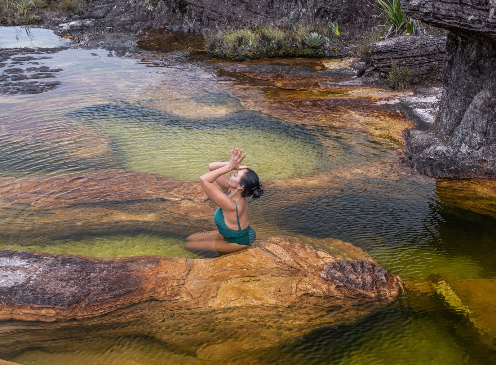 Un bassin naturel au sommet du monde, rien que pour soi. Une femme en maillot de bain vert sauge est agenouillée dans un bassin naturel peu profond sur le trek du Mont Roraima, les mains jointes au-dessus de la tête dans une posture méditative, entourée de roches aux teintes ocre