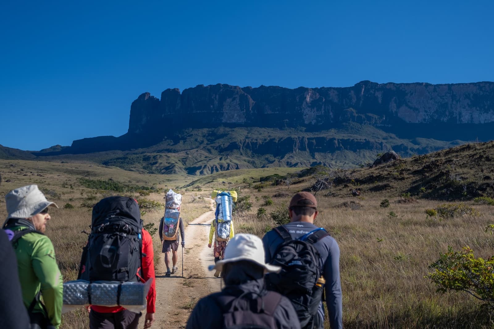 Le mont Roraima au bout du chemin, les jambes qui décident du reste. Un groupe de randonneurs portant de lourds sacs à dos sur le trek du mont Roraima, marchant sur un chemin de terre en savanne vers les falaises verticales du tepui sous un ciel bleu intense