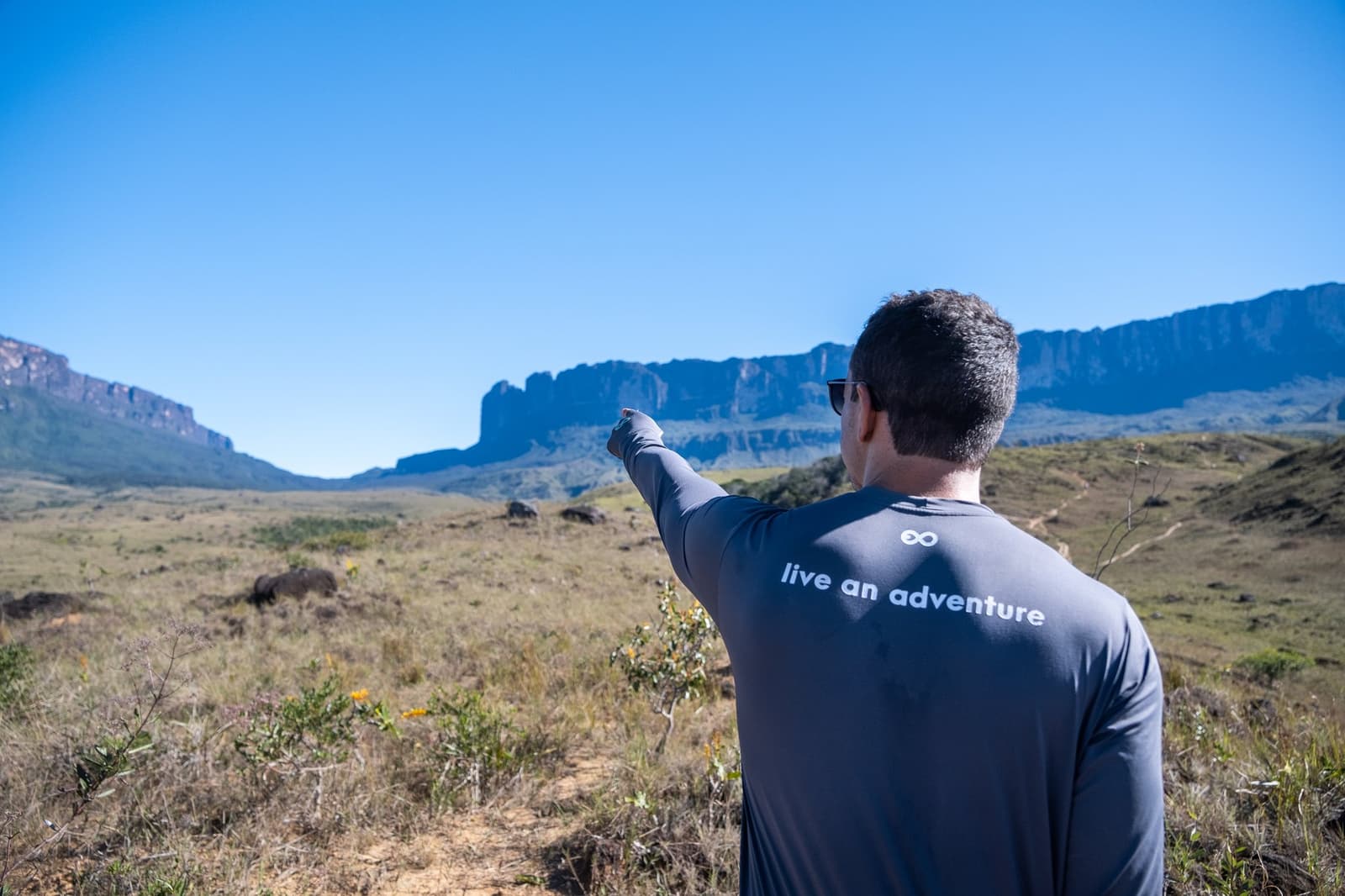 Voilà votre objectif. Ce qui vous attend là-haut, c'est à vous de le découvrir. Un guide portant un t-shirt 'live an adventure' désigne les falaises imposantes d'un tepui lors du trek du mont Roraima, avec une vaste savane s'étendant sous un ciel bleu limpide