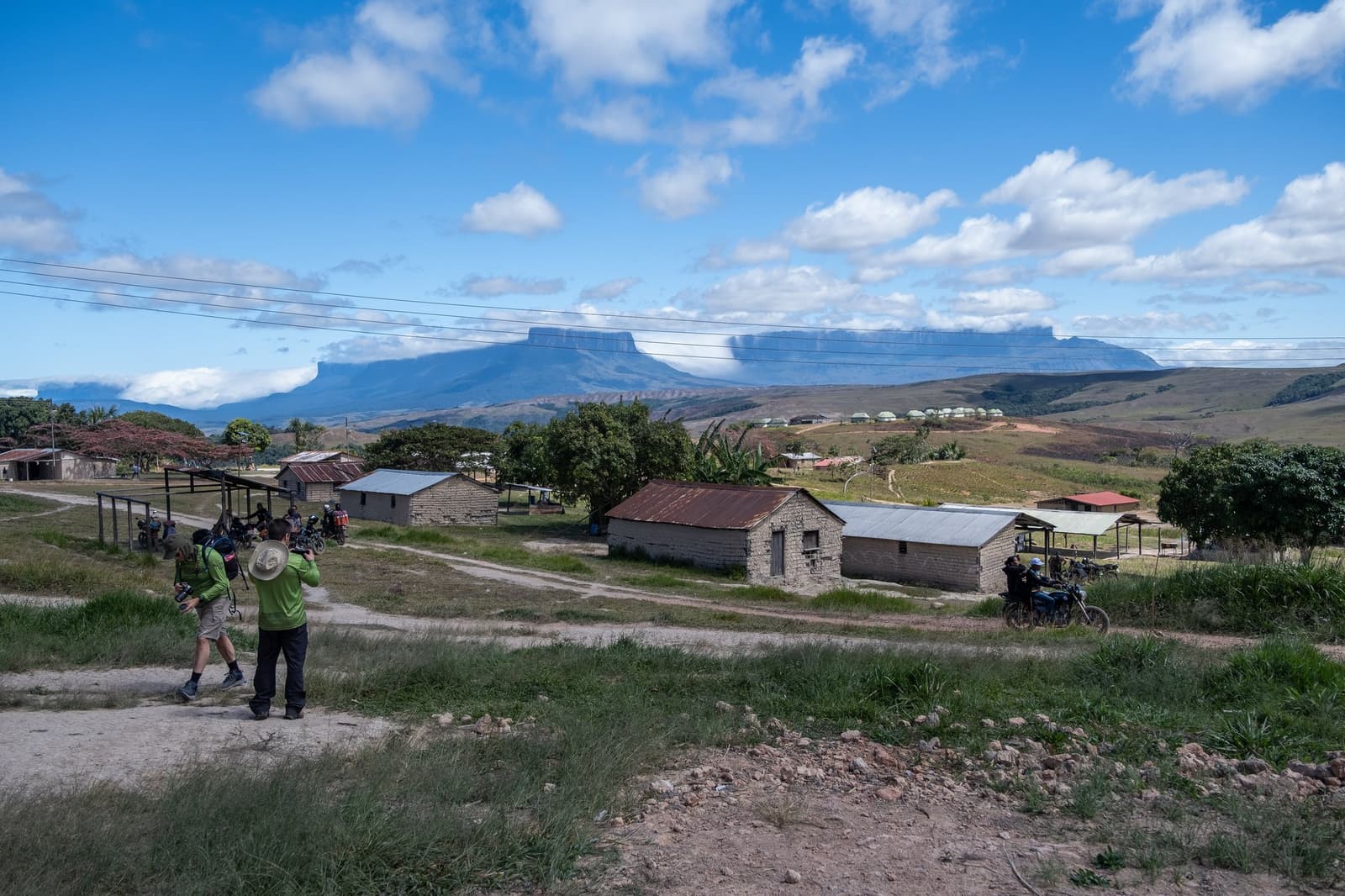 Le village vit à l'ombre d'un des plateaux les plus célèbres du monde. Deux photographes sur un chemin de terre au-dessus d'un petit village lors du trek du Mont Roraima, avec un tepui au sommet plat se découpant à l'horizon sous un ciel bleu