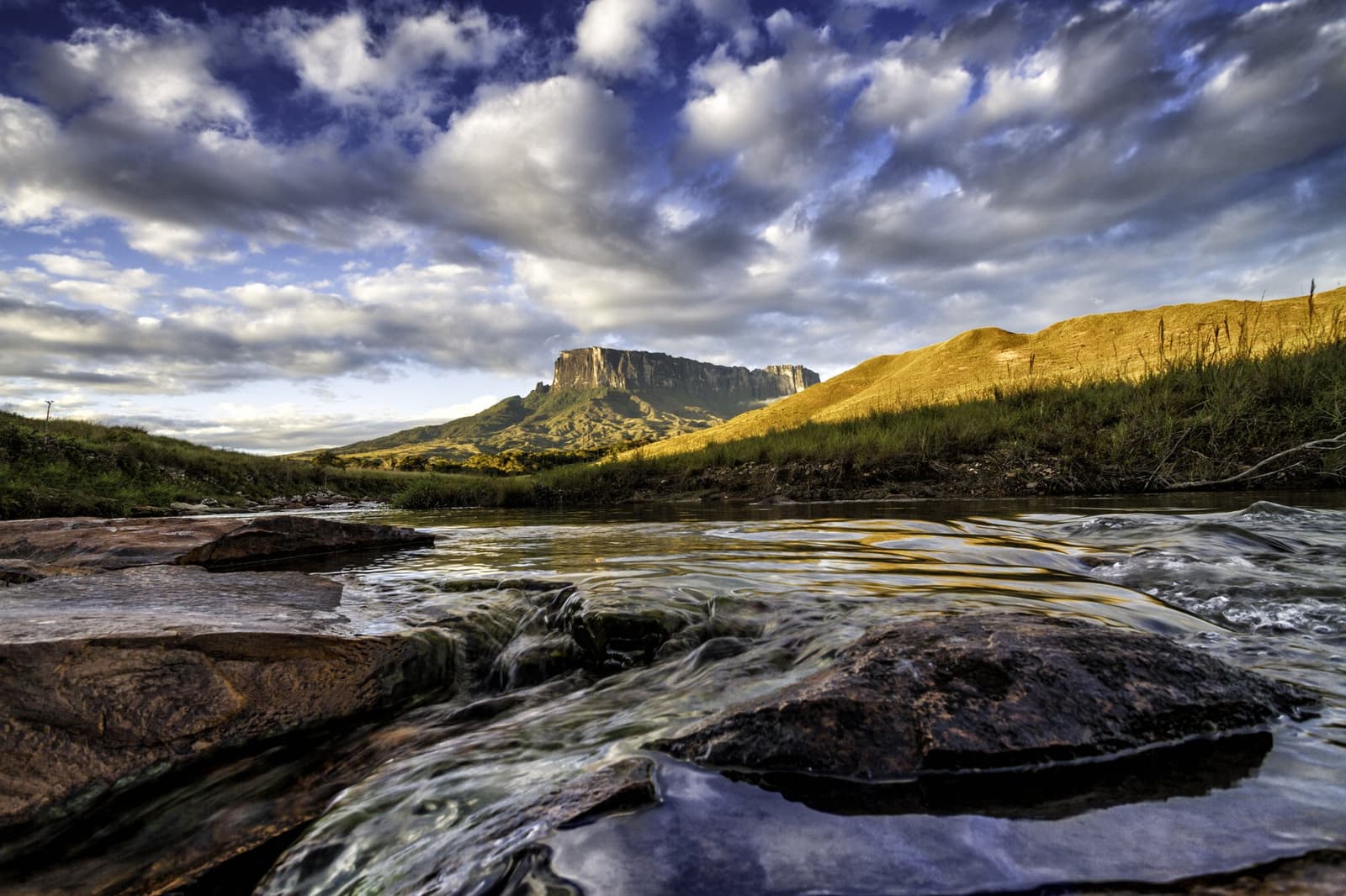 La rivière file vers le tepui, et vous la suivez. Une rivière peu profonde aux rochers parsemés coule vers le sommet tabulaire du mont Roraima sous un ciel dramatique lors du trek du mont Roraima