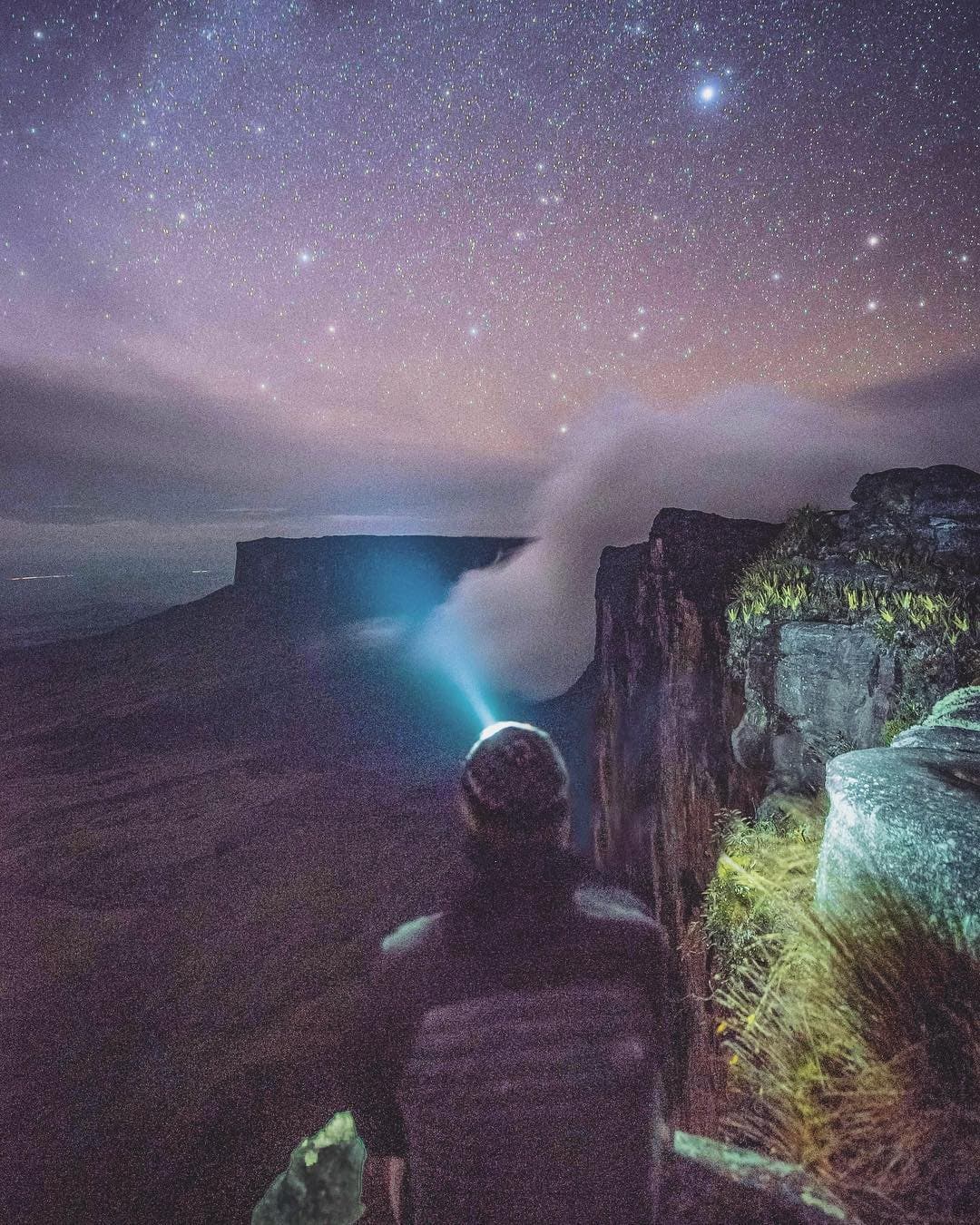 À cette altitude, on ne regarde plus le ciel, on l'habite. Un randonneur solitaire équipé d'une lampe frontale se tient au bord d'une falaise de tepui lors du trek du mont Roraima, contemplant une mer de brume vers un plateau lointain sous un ciel étoilé