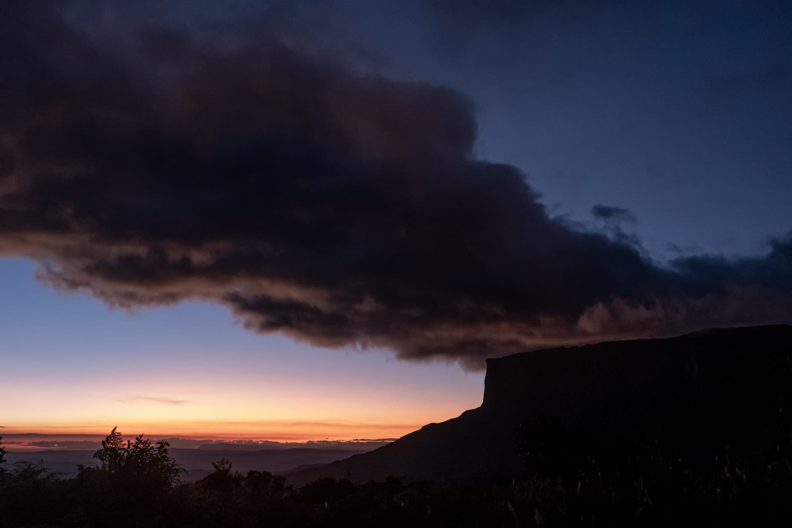 La montagne choisit l'ambiance, et ce soir elle a opté pour le dramatique. Un nuage d'orage spectaculaire s'étend au-dessus de la silhouette d'un tepui lors d'un coucher de soleil flamboyant sur le trek du mont Roraima, avec un ciel orange et bleu profond à l'horizon