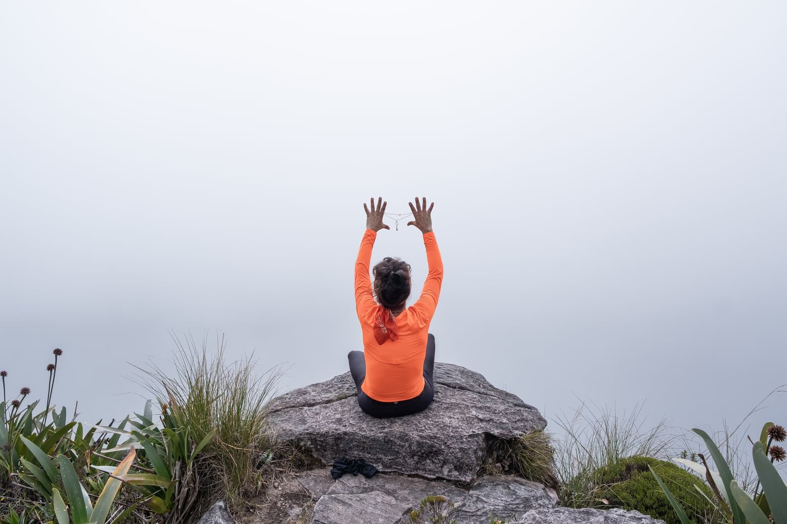 Quand le panorama disparaît dans le brouillard, on lève les bras et on s'en fiche. Une femme en haut orange est assise en tailleur sur un rocher moussu au sommet lors du trek du mont Roraima, les bras levés vers un ciel noyé dans un épais nuage blanc, entourée de broméliacées des hautes terres