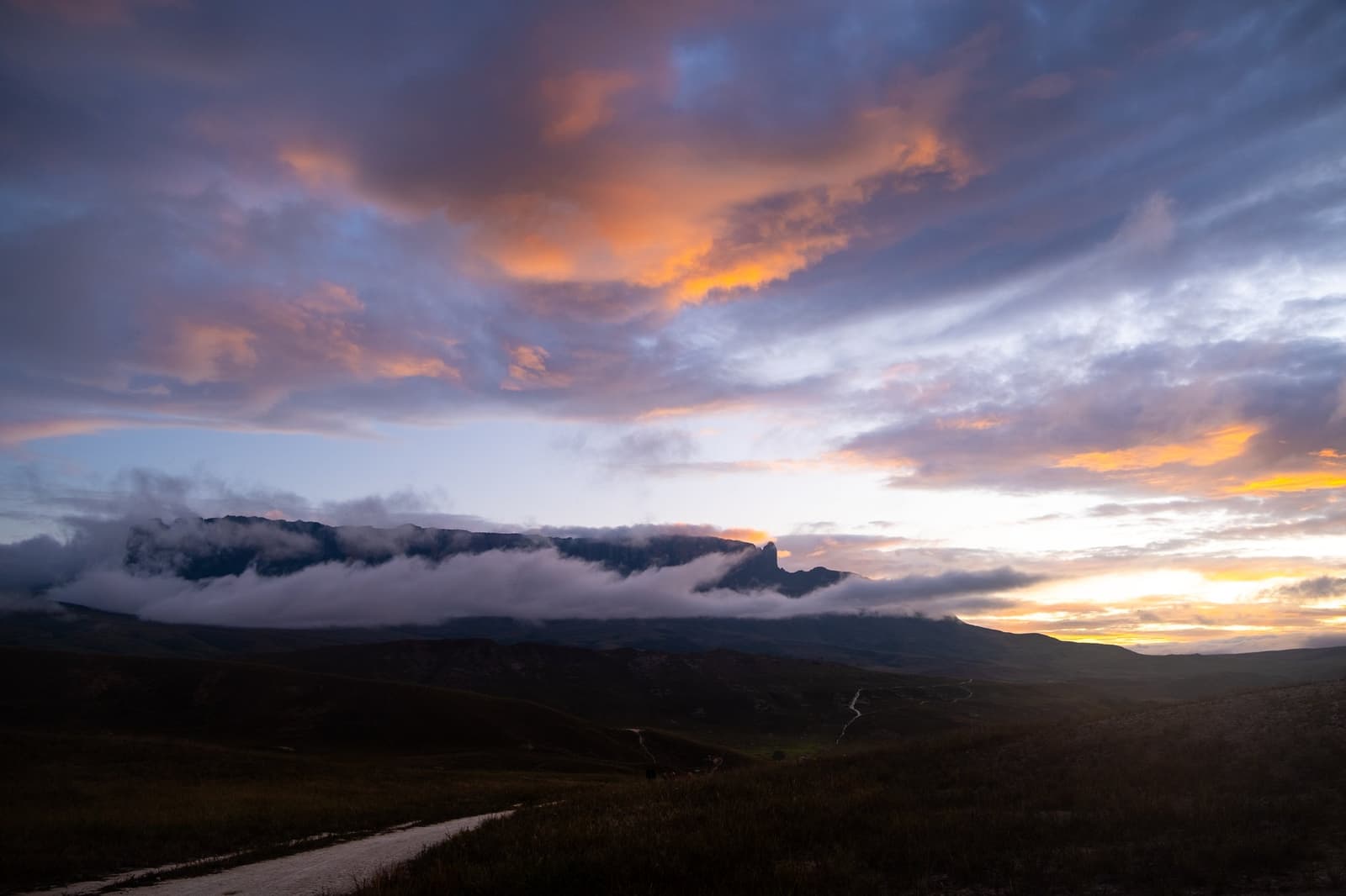 La lumière du soir ici ne ressemble à rien d'autre. Un ciel de coucher de soleil spectaculaire en orange et violet plane sur la silhouette d'un tepui à sommet plat noyé dans les nuages lors d'un trek au mont Roraima, avec un sentier sinueux visible dans la savane sombre au premier plan