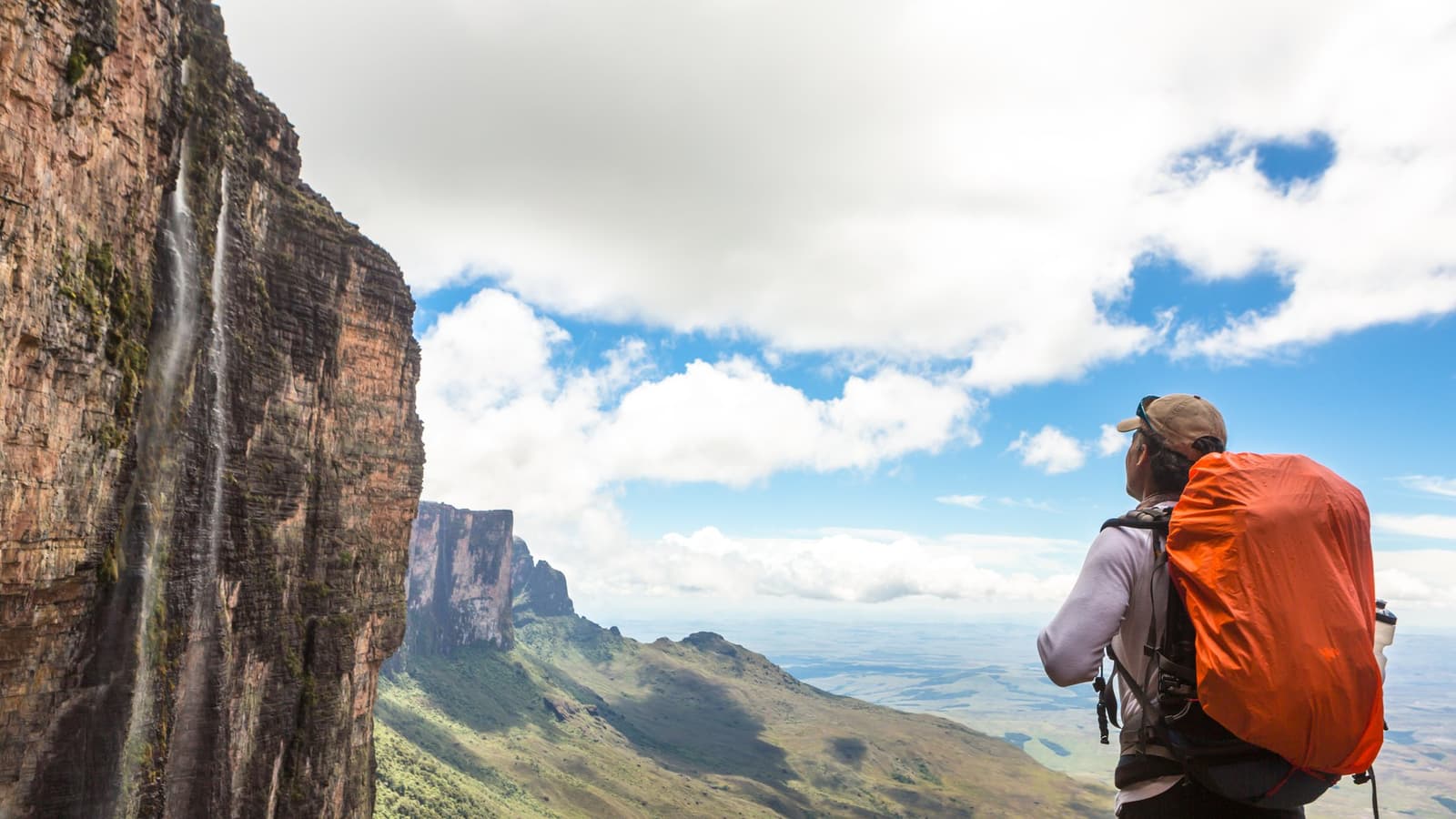 Il y a des panoramas qui font oublier le poids du sac. Un randonneur portant un sac à dos orange se tient au bord d'une imposante paroi de grès sur le trek du Mont Roraima, contemplant un vaste paysage de savane pendant qu'une fine cascade dévale la falaise verticale à ses côtés